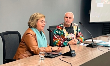 Uma mulher e um homem estão sentados em um palco fazendo uma palestra