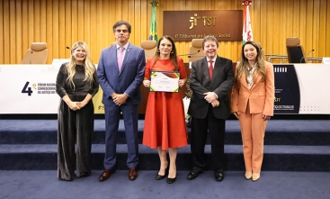 Fotografia na qual cinco pessoas posam em auditório do TST; ao centro, mulher segura certificado