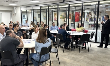 Grupo de pessoas participa de palestra em ambiente corporativo, com mesas redondas e obras de arte expostas ao fundo.