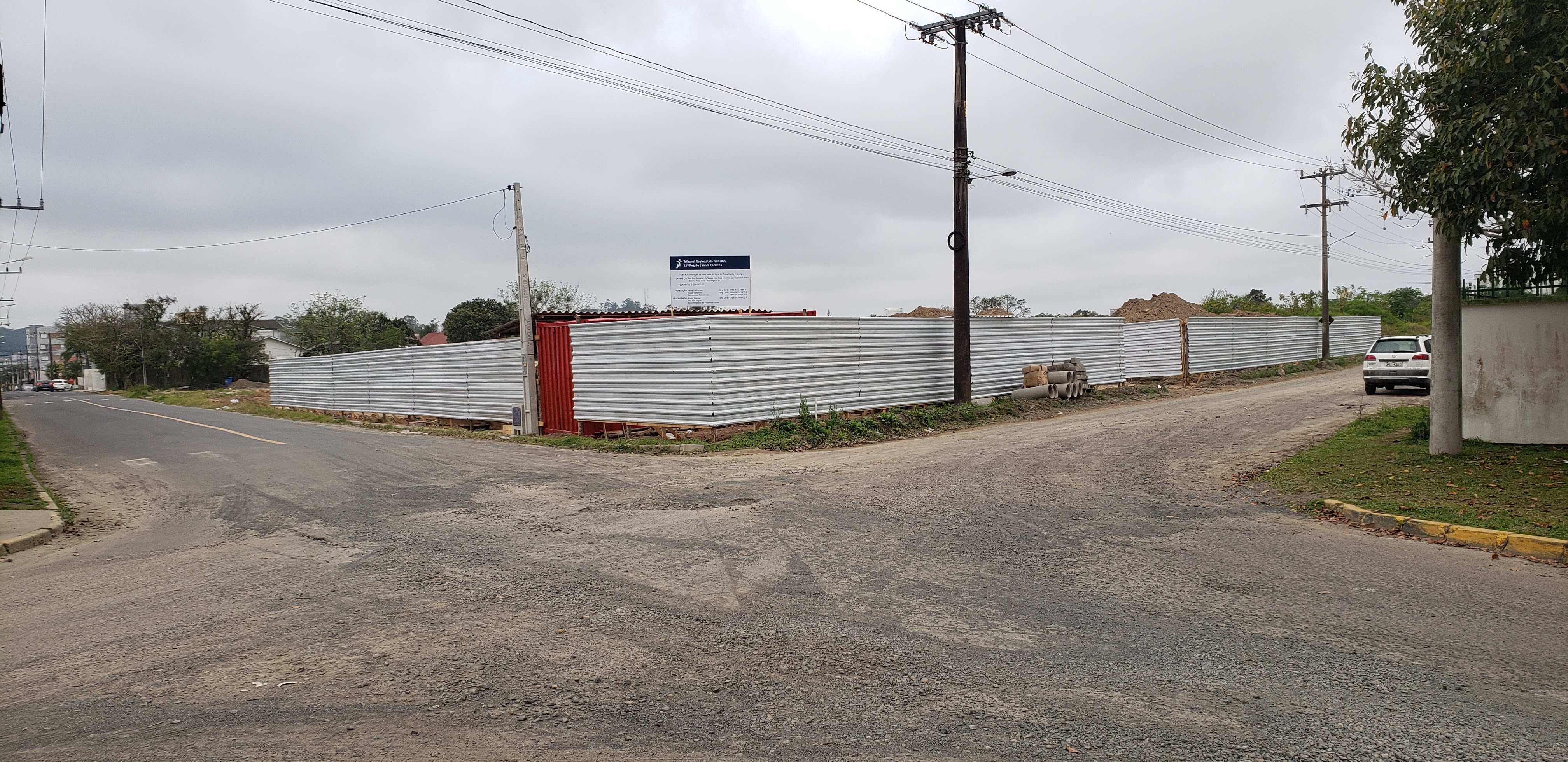 vista geral da esquina do terreno cercado com tapume em telha metálica, placa da obra fixada acima do tapume