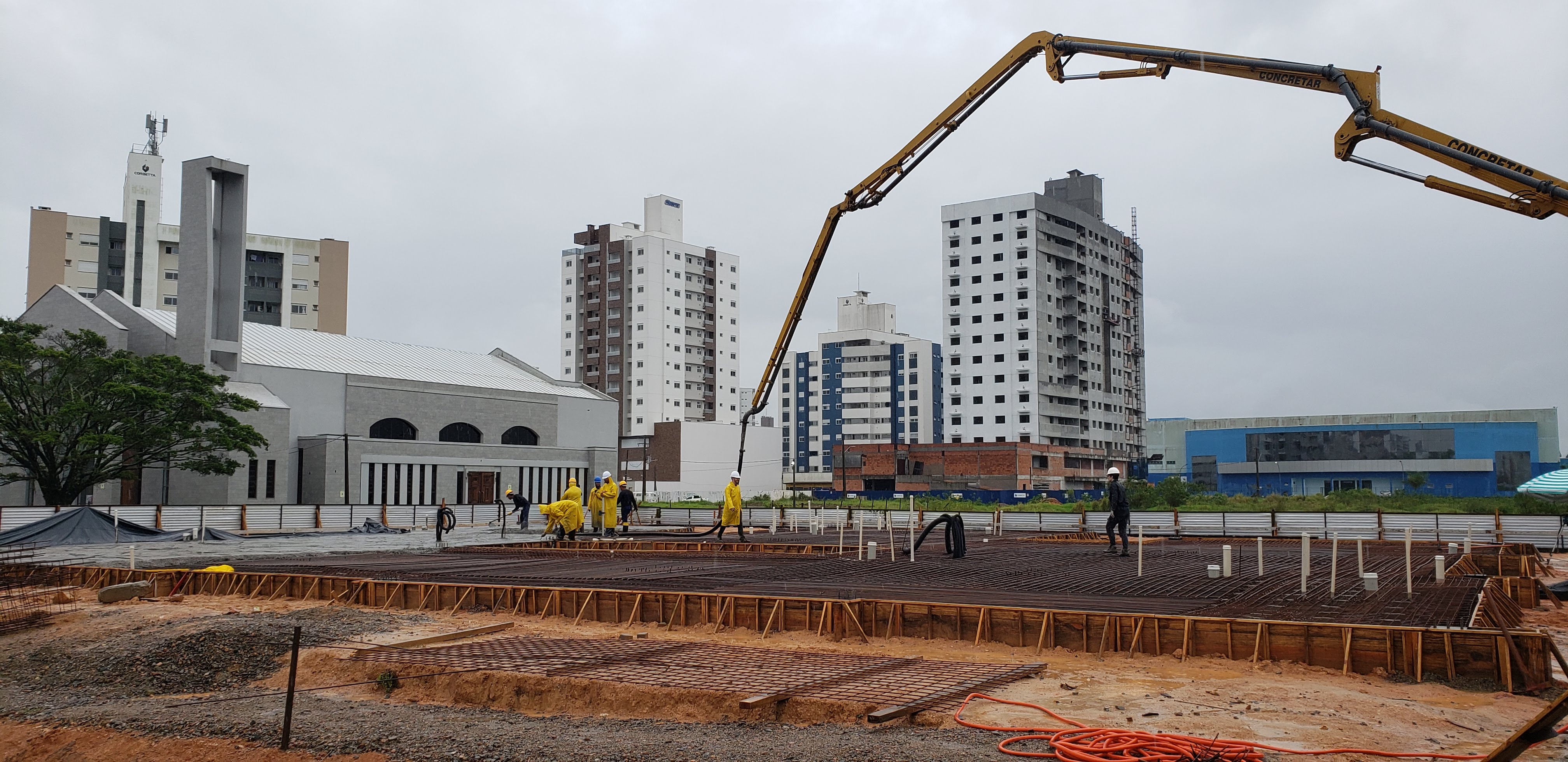 Foto - dia chuvoso, com operários sobre estrutura em ferro trabalhando na concretagem do radier, no alto lança do caminhão de concreto.