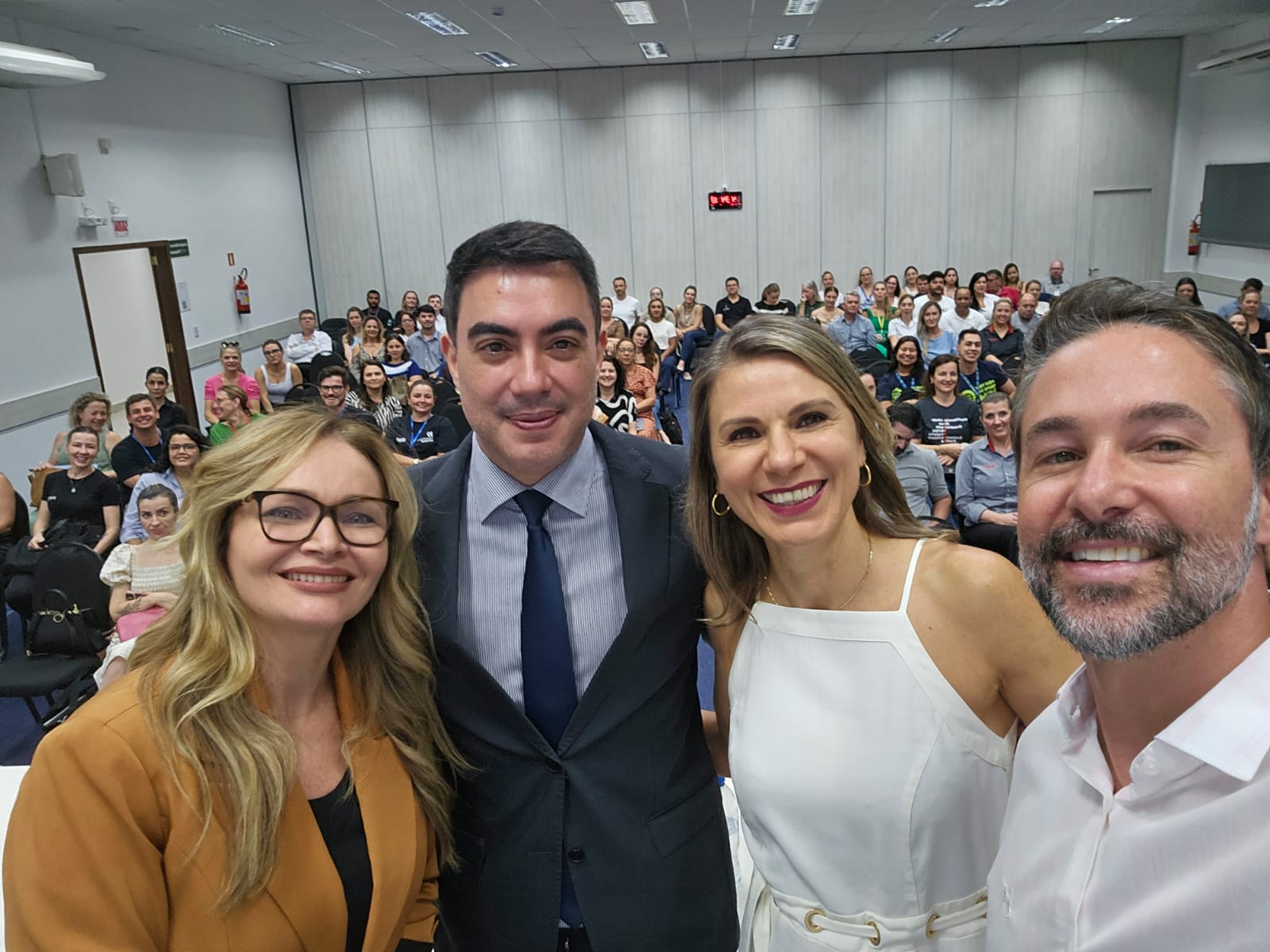 Quatro pessoas estão na frente, sorrindo para uma selfie, com um auditório cheio ao fundo. A imagem transmite um clima alegre e participativo, durante a audiência pública