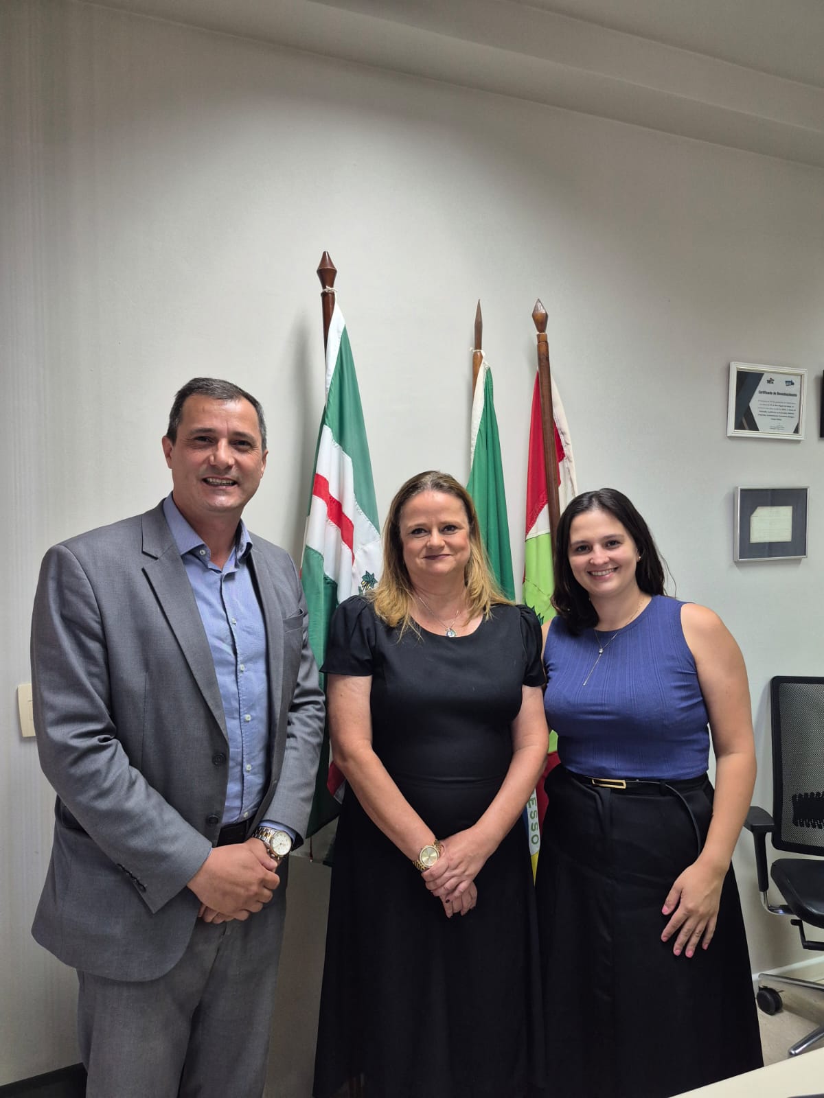  Juíza Ana Rick e membros da OAB posam para foto em frente à uma parede branca, com bandeiras ao fundo