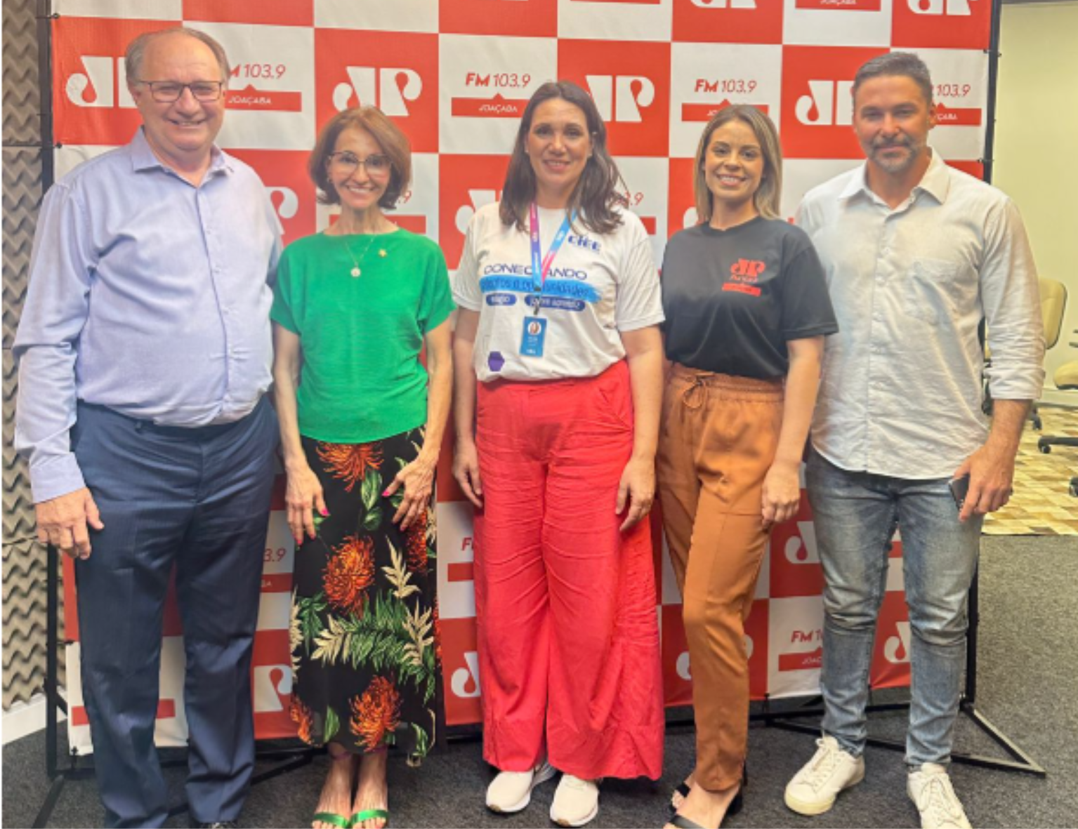 Cinco pessoas estão em pé, lado a lado, posando para a foto em frente a um painel com a logomarca da rádio Jovem Pan e a inscrição “FM 103.9 Joaçaba”. Da esquerda para a direita,  Sr. Luis Carlos Floriani, Juíza Lisiane Vieira, Lilian Henn (Coordedora do CIEE de Joaçaba), Elisandra Costa (Repórter da Joven Pan/Rádio Líder Herval D Oeste) e Cristian Santiani (Gerente do CIEE  - SC). Todos sorriem para a câmera em ambiente de estúdio.