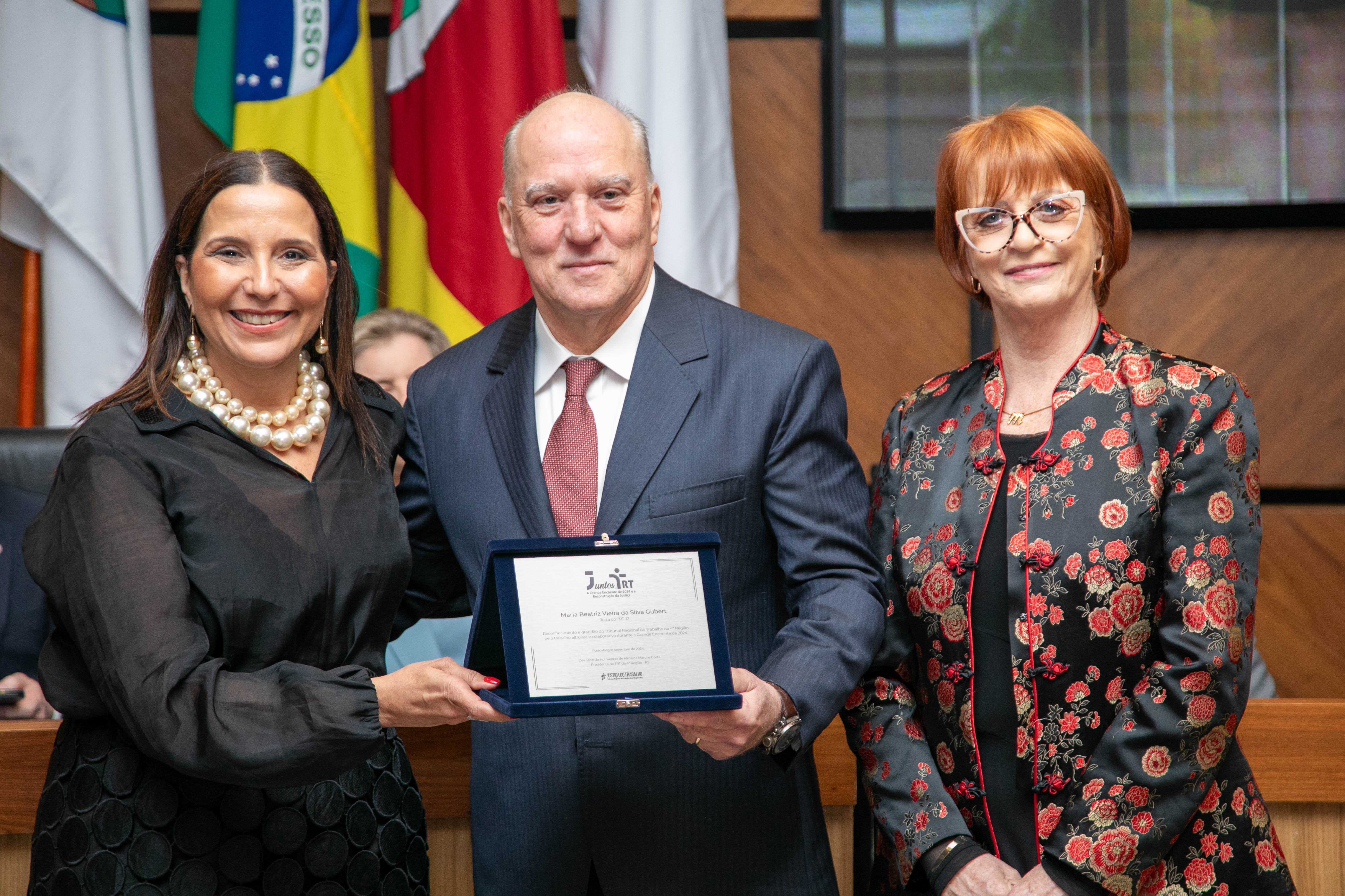 Fotografia de duas mulheres e um homem vestidos formalmente. Eles sorriem para a câmera. Uma das mulheres e o homem seguram uma placa de metal.