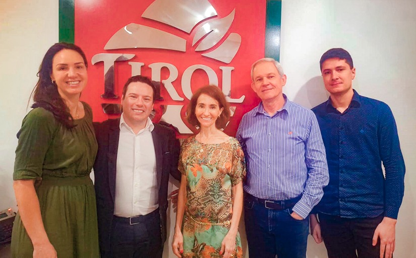 A foto mostra cinco pessoas em pé, sorrindo, posando diante de um painel com o logotipo da empresa "Tirol" em destaque. As pessoas estão vestidas de maneira casual, mas arrumadas, indicando um ambiente corporativo ou de visita institucional. O grupo inclui duas mulheres e três homens