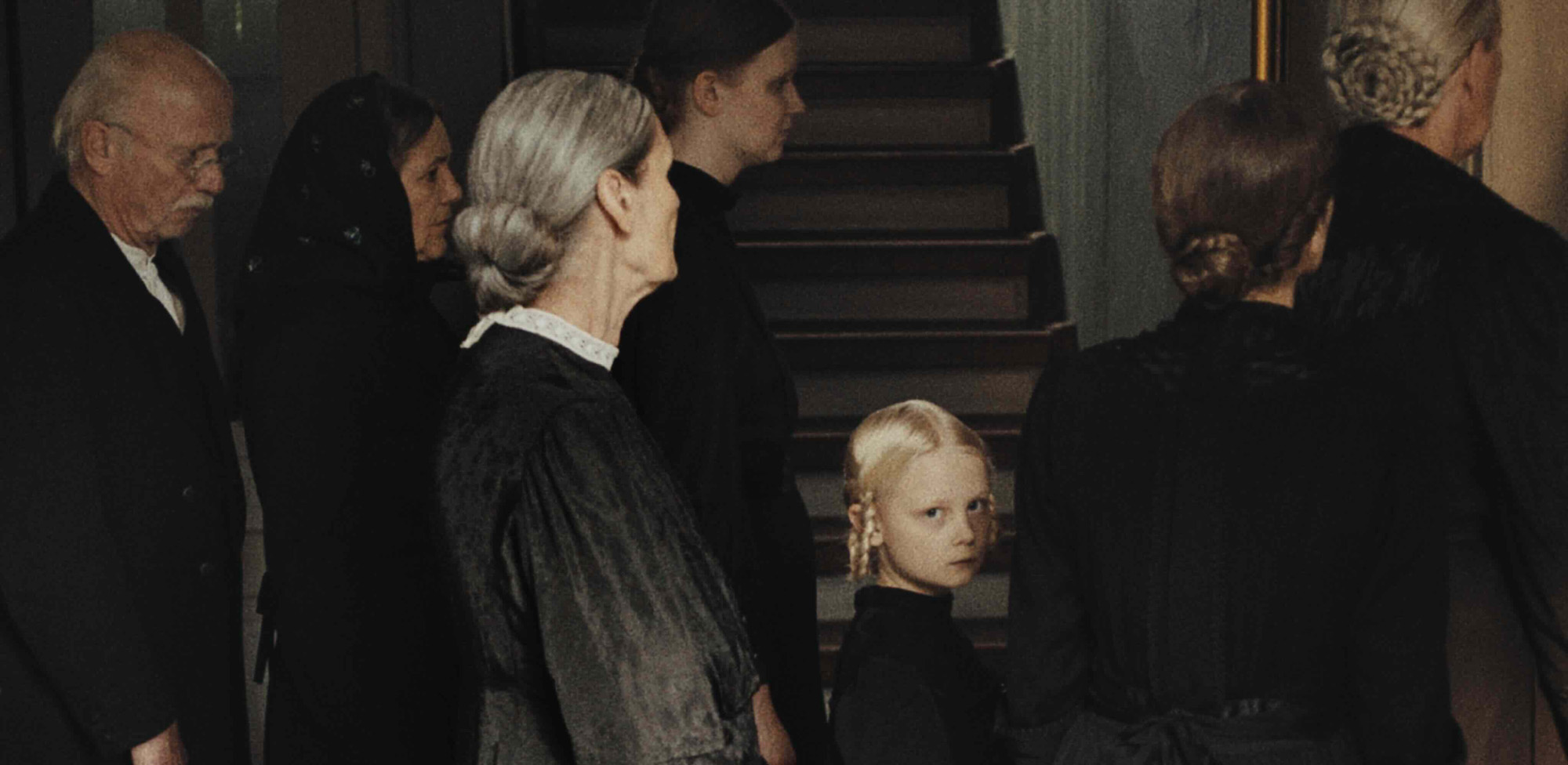 Para todos verem: Banner em que há cinco mulheres, um senhor e uma menina vestidos de preto. A menina está olhando para frente. Ao fundo há uma escada.