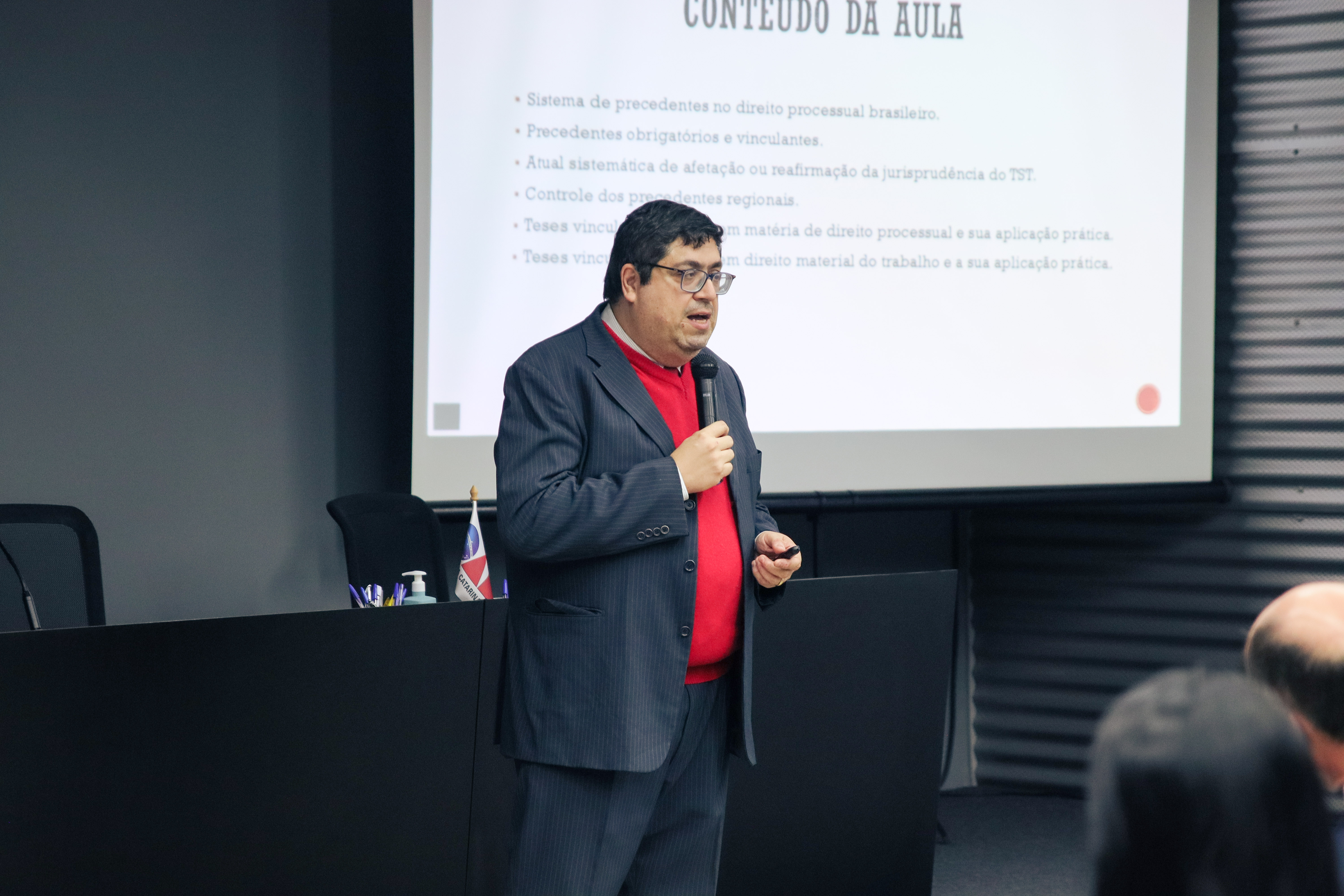Foto de um homem vestindo terno cinza e blusa vermelha. Ele fala ao microfone