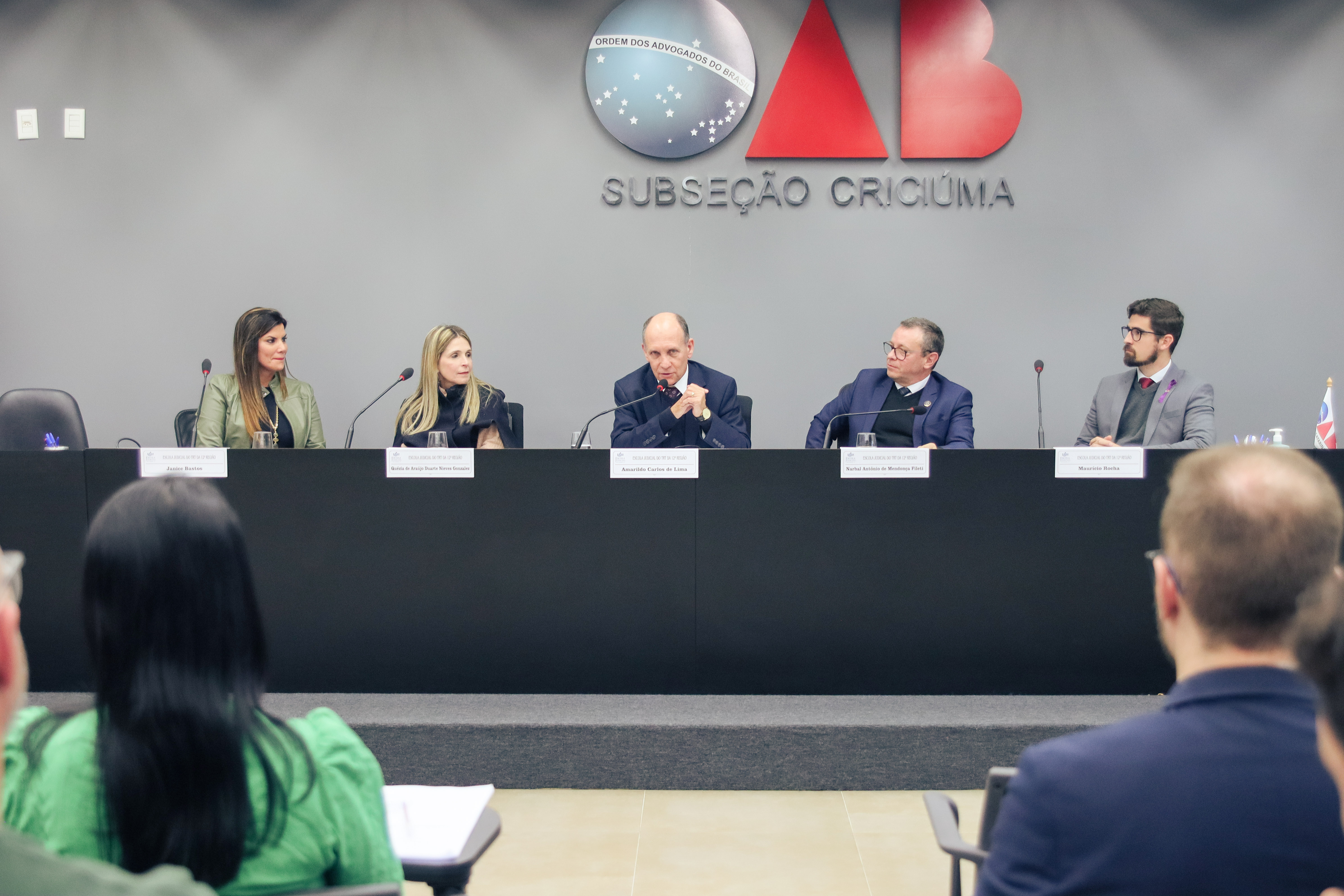 Foto de uma mesa de honra. Ao centro, um homem fala ao microfone