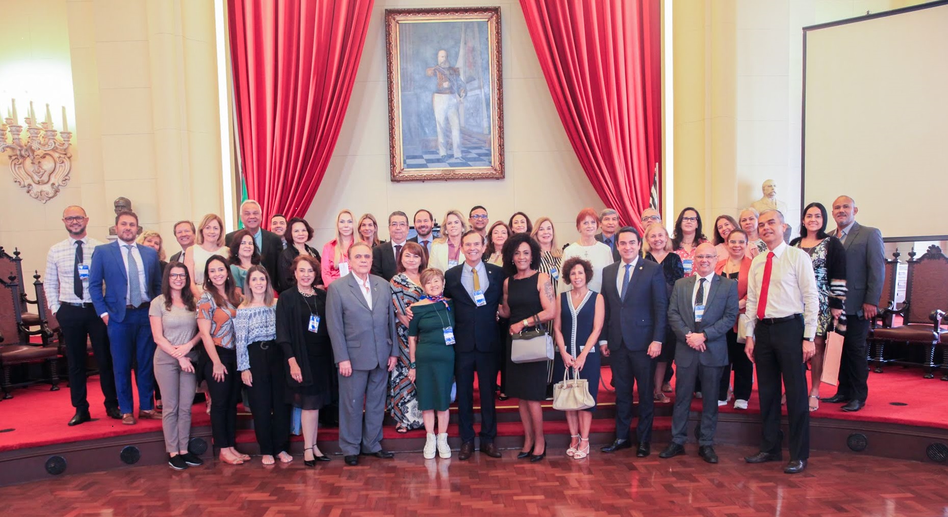 Grupo de cerca de 40 pessoas posam para foto em um salão, com piso de tacos de madeira lustrada, cortinas vermelhas ao fundo e um quadro de um oficial do império