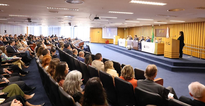 Foto aberta mostra um auditório amplo, com uma mesa de autoridades e uma mulher em um púlpito à direita, além de cerca de 100 pessoas sentadas