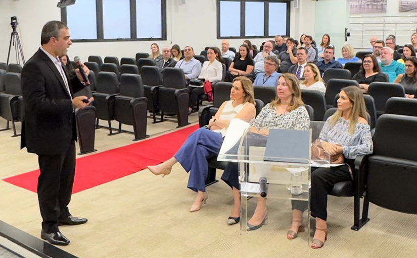 Palestrante fala ao público em auditório, enquanto espectadores atentos acompanham a apresentação sentados nas poltronas.