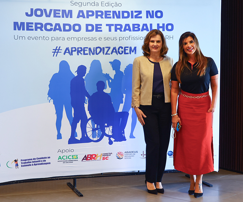 Duas mulheres posam diante de painel do evento “Jovem Aprendiz no Mercado de Trabalho”, em ambiente corporativo.