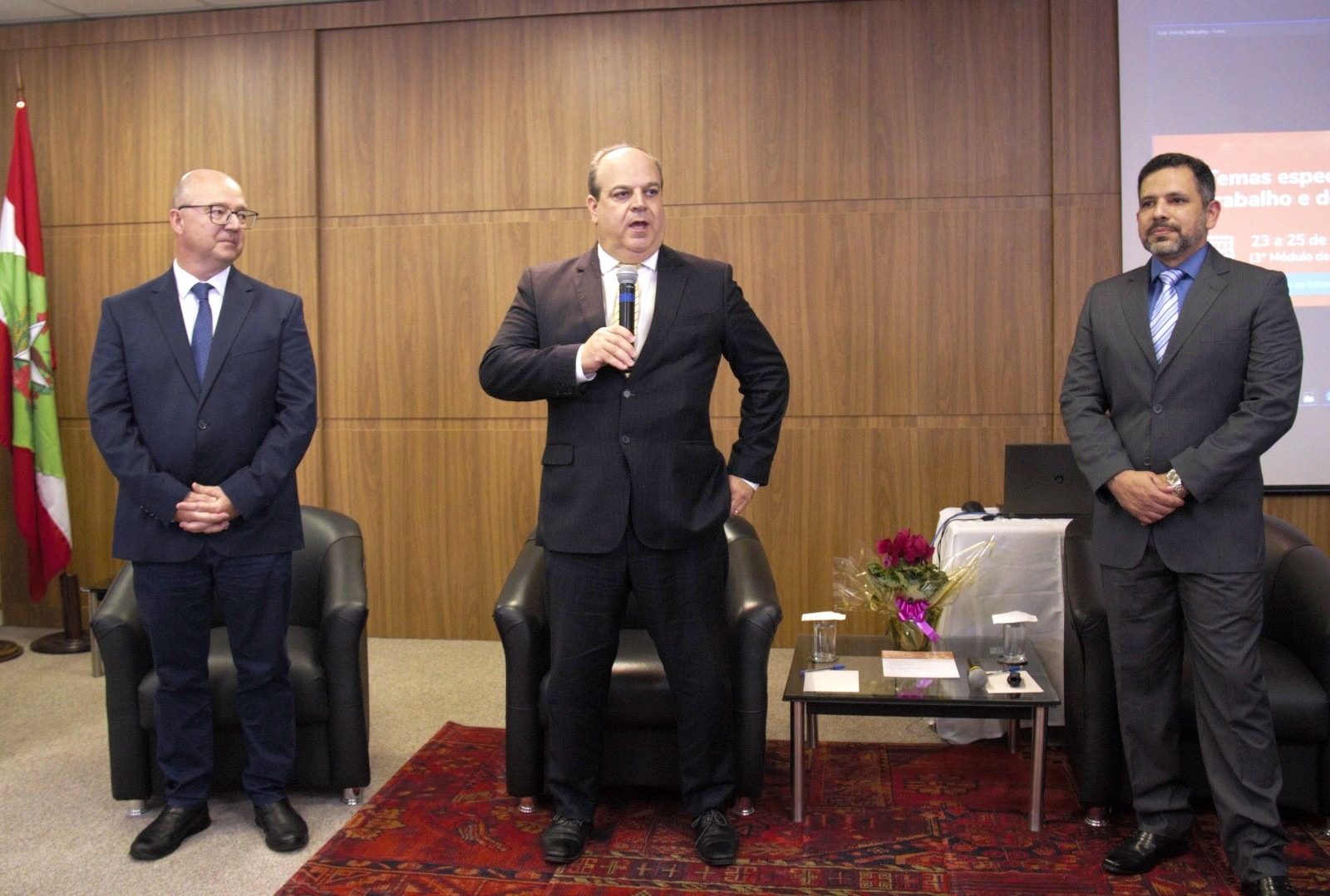 Três homens de terno e gravata em pé, na frente de um auditório. O homem do meio está segurando um microfone e falando.