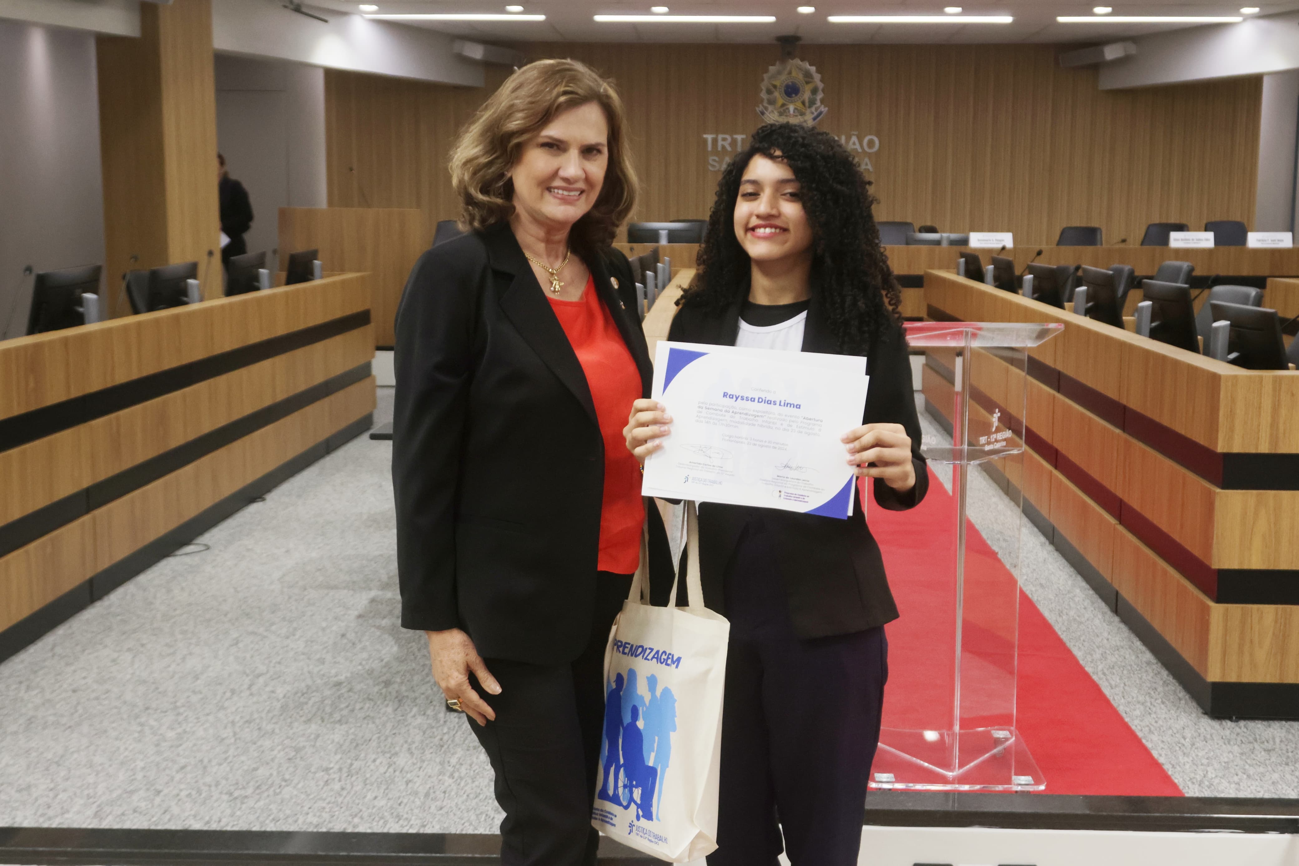 Foto mostra uma mulher madura, loira, vestida formalmente, e uma jovem de cerca de 16 anos, de pele morena e cabelos cacheados negros, que segura um certificado. Ambas estão sorridentes