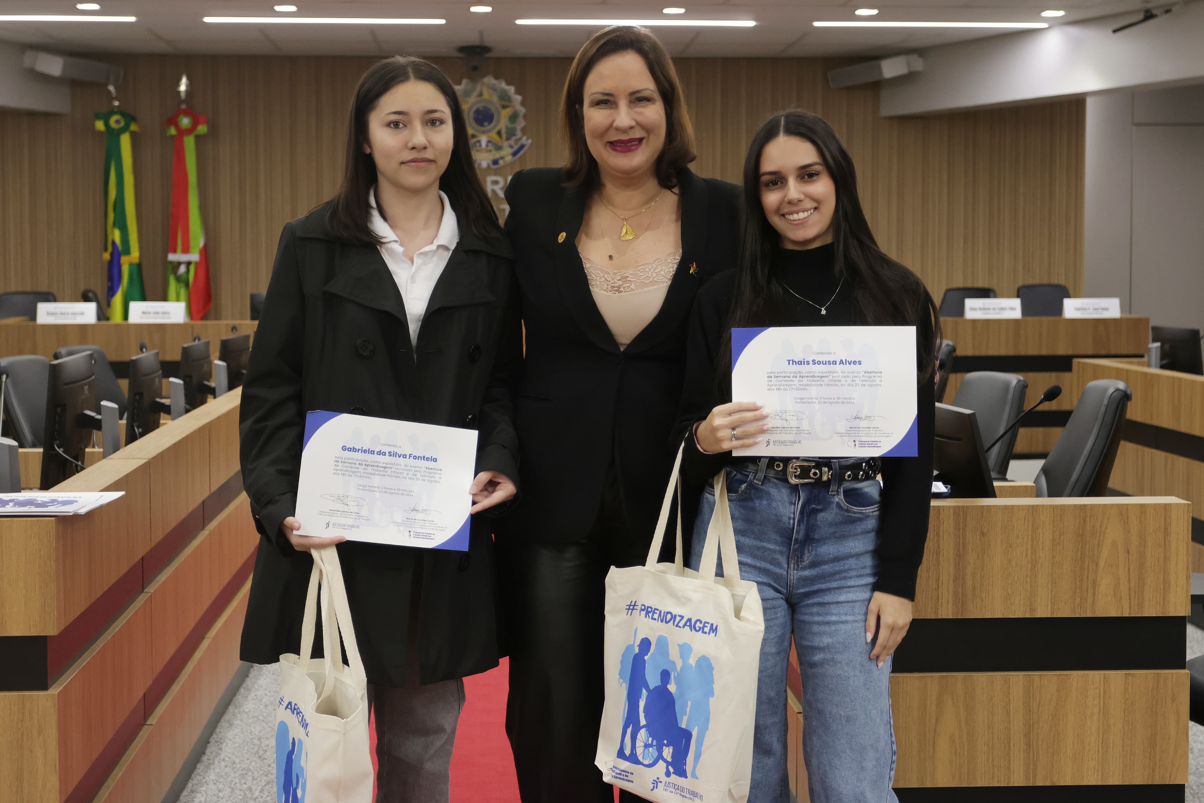 Foto mostra uma mulher de cerca de 50 anos, pele branca e cabelos castanhos, ao lado de duas jovens de cerca de 18 anos, ambas de pele branca e cabelos lisos escuros. As jovens seguram certificados. As três estão sorridentes.