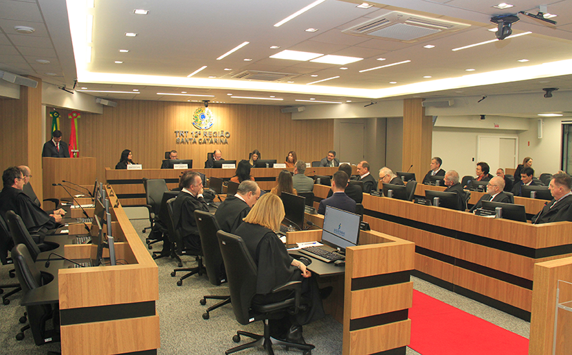 Fotografia da sala de sessões do Tribunal Pleno durante a solenidade. Ao centro cinco pessoas sentadas olham para a mesa diretiva que tem seis integrantes, sendo três mulheres e três homens. 