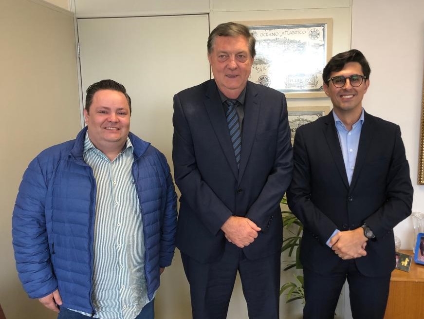 Três homens estão de pé, dois de terno e gravata e outro de jaqueta azul. Estão sorrindo e posando para a foto.