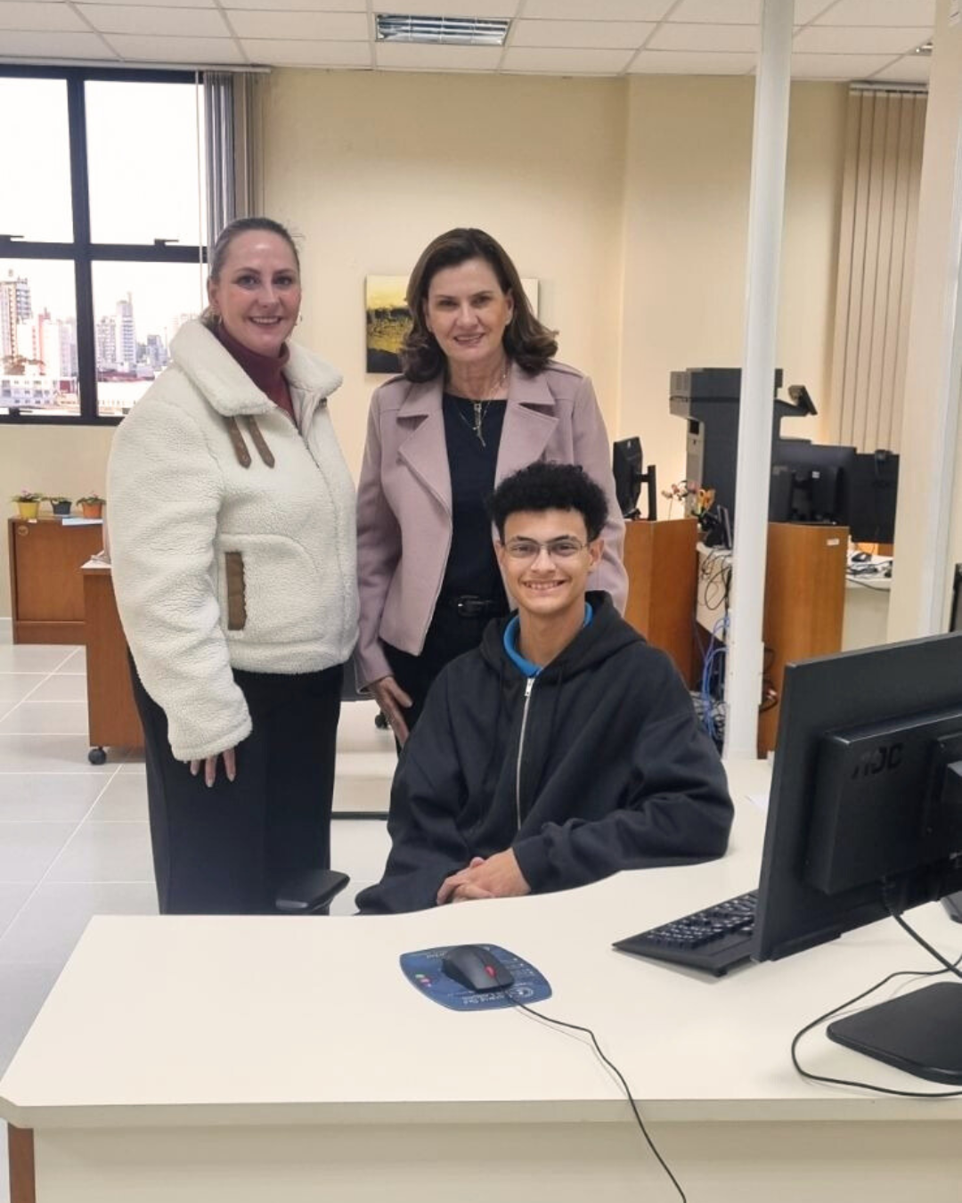 Duas mulheres estão em pé e um jovem rapaz está sentado. Os três estão sorrindo para a foto