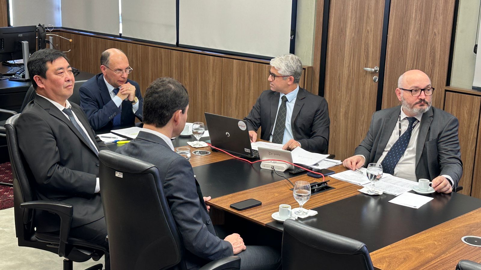 Foto lateral de um grupo de cinco homens de terno escuro, ao redor de uma mesa retangular, olhando para um telão. 