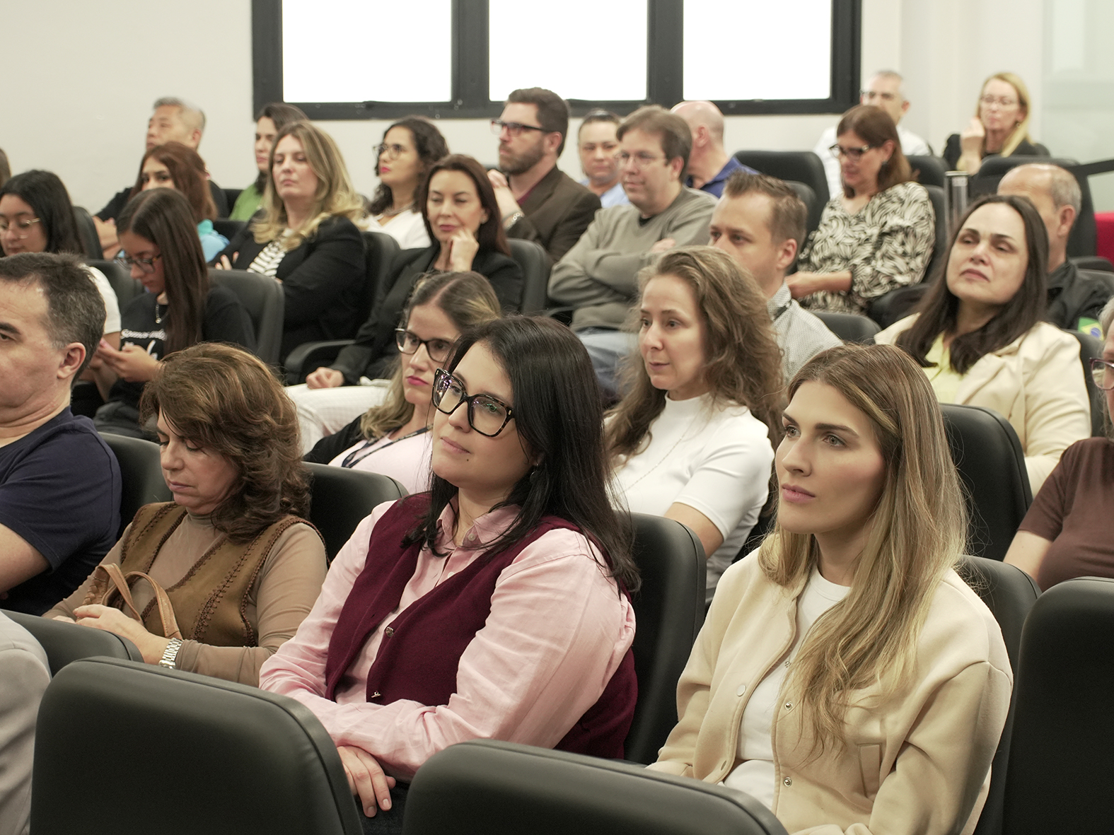 Cerca de 30 pessoas acompanham sentados alguém falando. Foto em ângulo de 45 graus