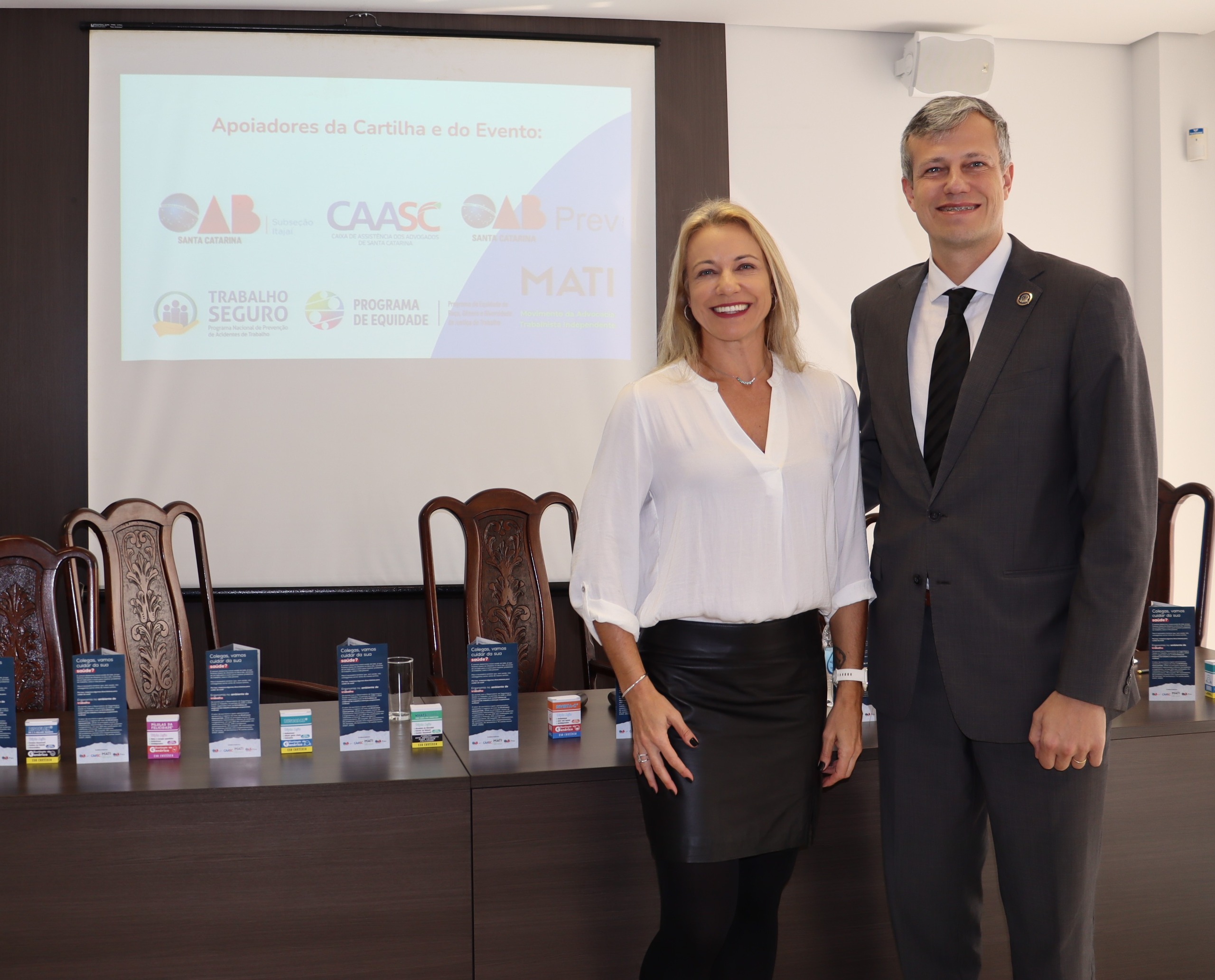 Foto de duas pessoas em pé em frente a uma mesa com caixas e panfletos. à esquerda, uma mulher loira, de camisa branca e saia preta e ao seu lado direito, um homem grisalho, alto, de terno preto