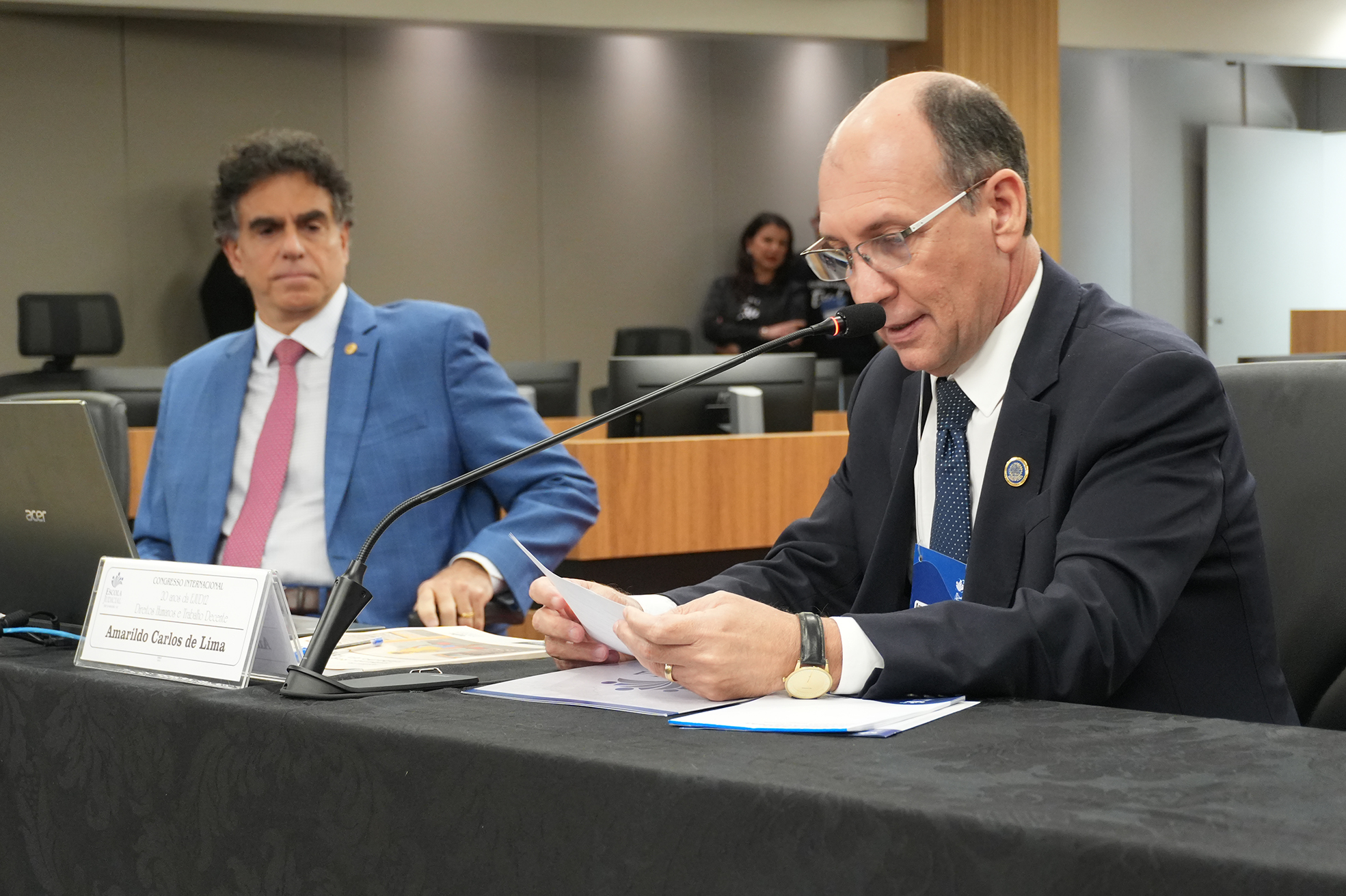 Foto de um homem de terno e gravata, usando óculos, falando no microfone sentado. Ao lado dele, outro homem de terno e gravata assistindo
