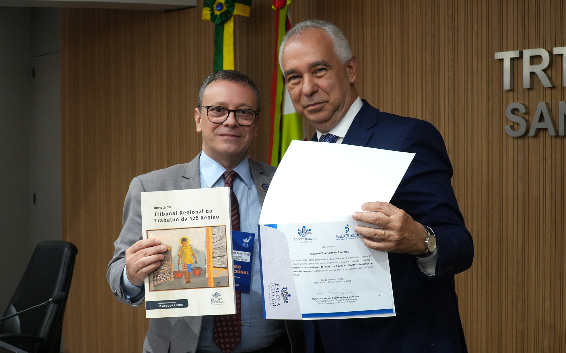 Dois homens de terno e gravata estão posando para a foto, segurando um livro e um certificado