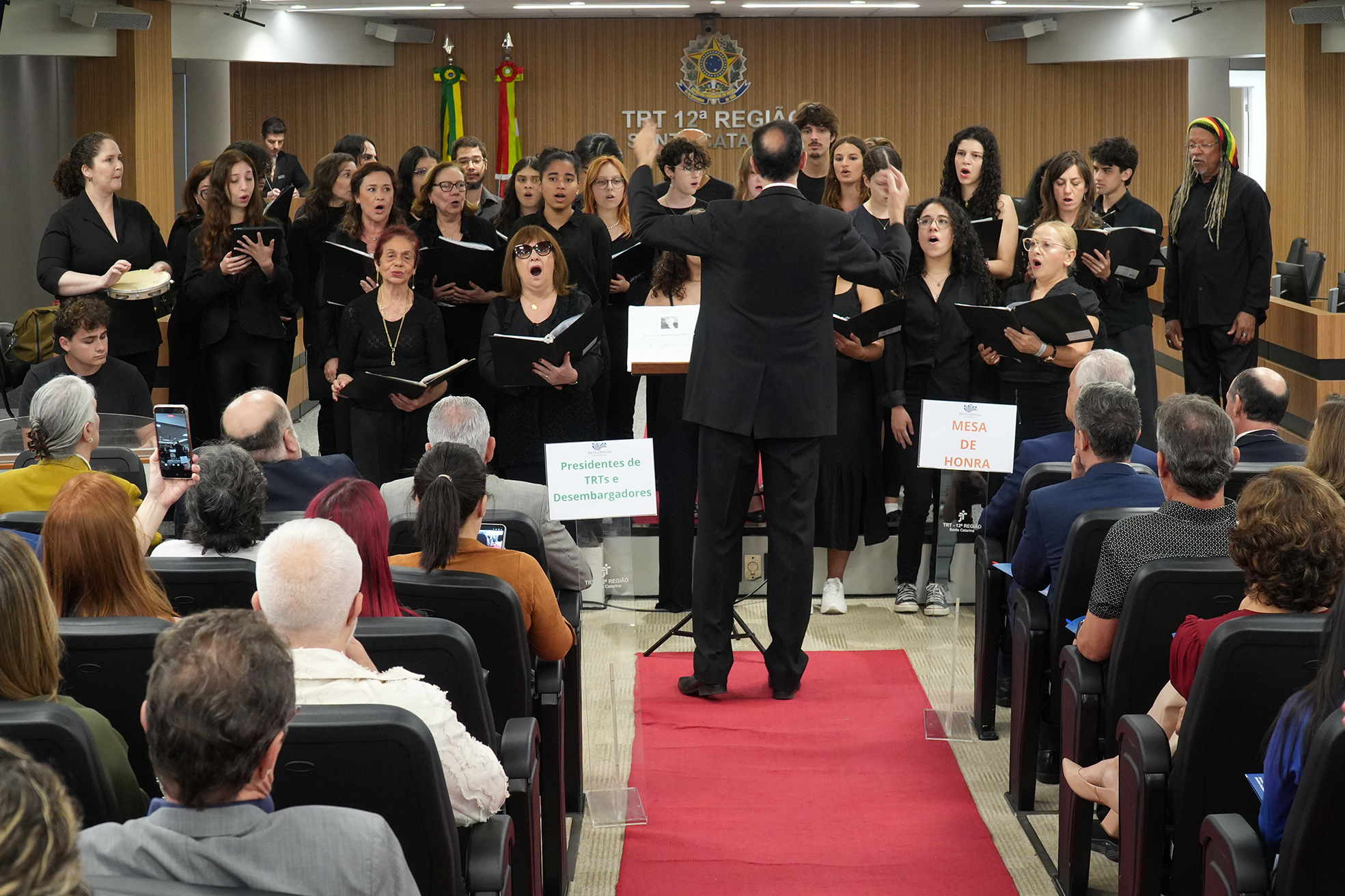 Foto de um coral de homens e mulheres cantando, vestidos de preto, durante um evento