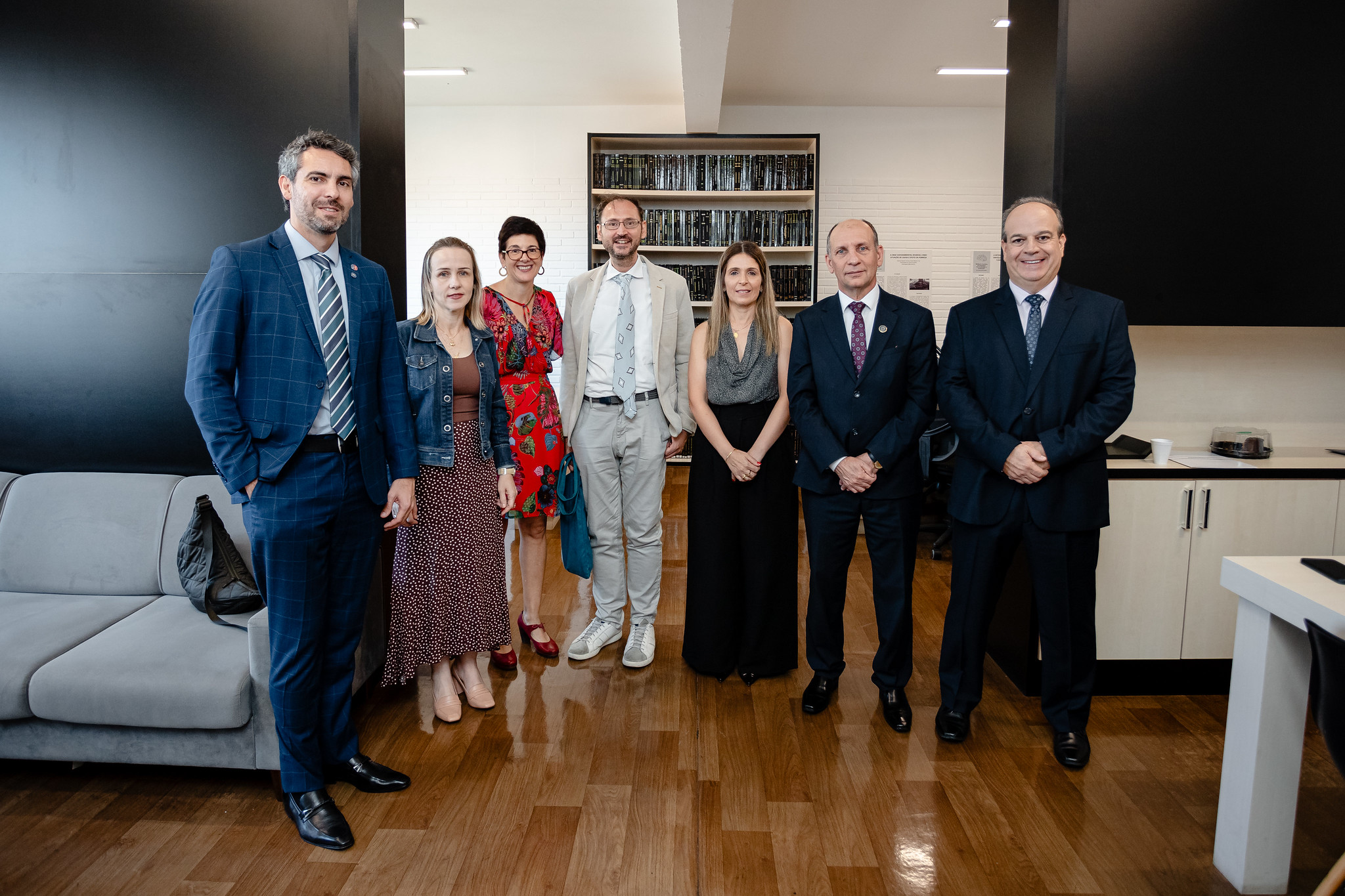 Quatro homens de terno e gravata e três mulheres estão em pé, posando para a foto em uma sala de reunião