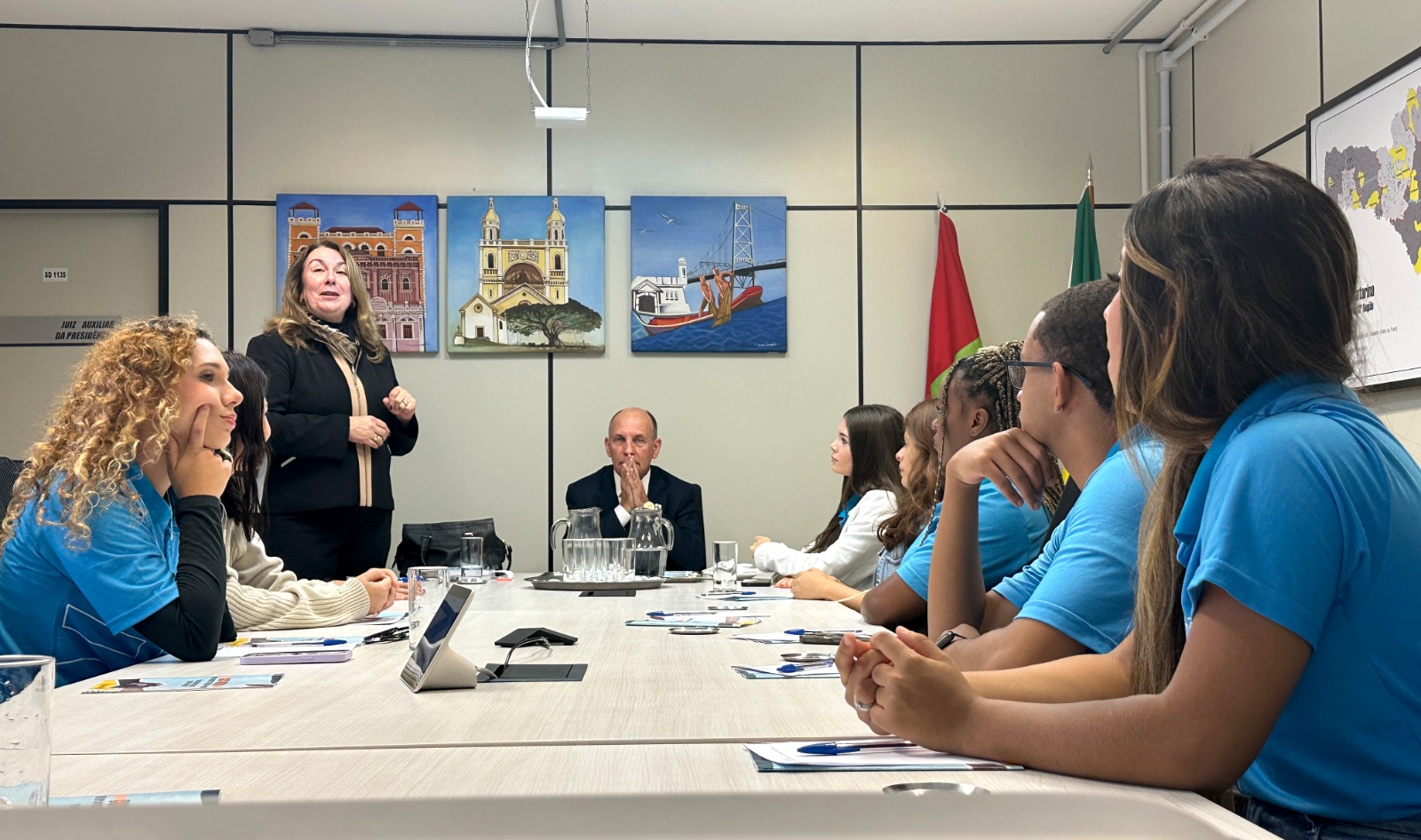 Jovens aprendizes estão sentados em uma mesa de reunião, na cabeceira está um homem de terno sentado e ao lado dele uma mulher loira em pé falando