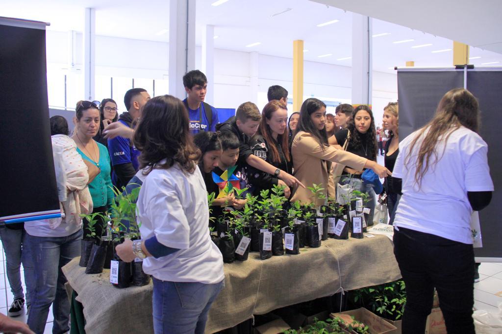 Fotografia de várias pessoas, entre eles jovens e adultos, estão em frente a uma mesa escolhendo mudas de plantas