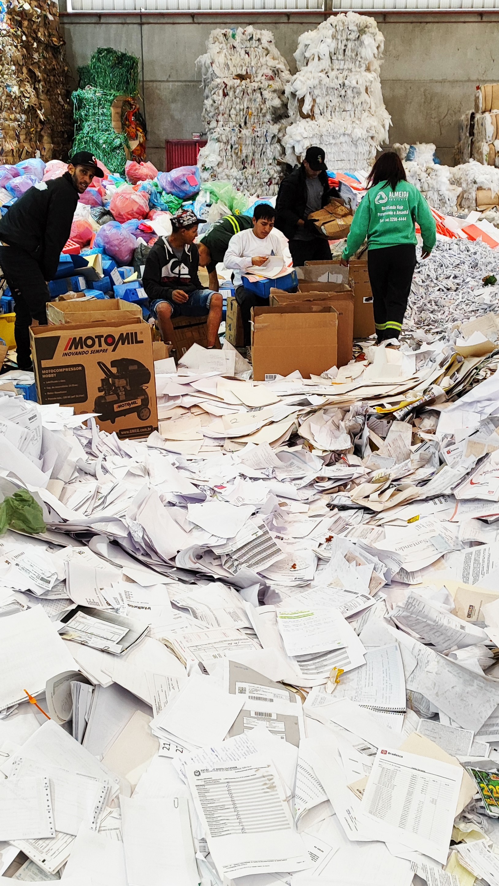 Foto vertical onde mais de metade da parte inferior é ocupada por papéis jogados no chão. Mais acima se vê seis pessoas manuseando os papéis e mais pilhas de papéis ao fundo do ambiente que lembra um armazém