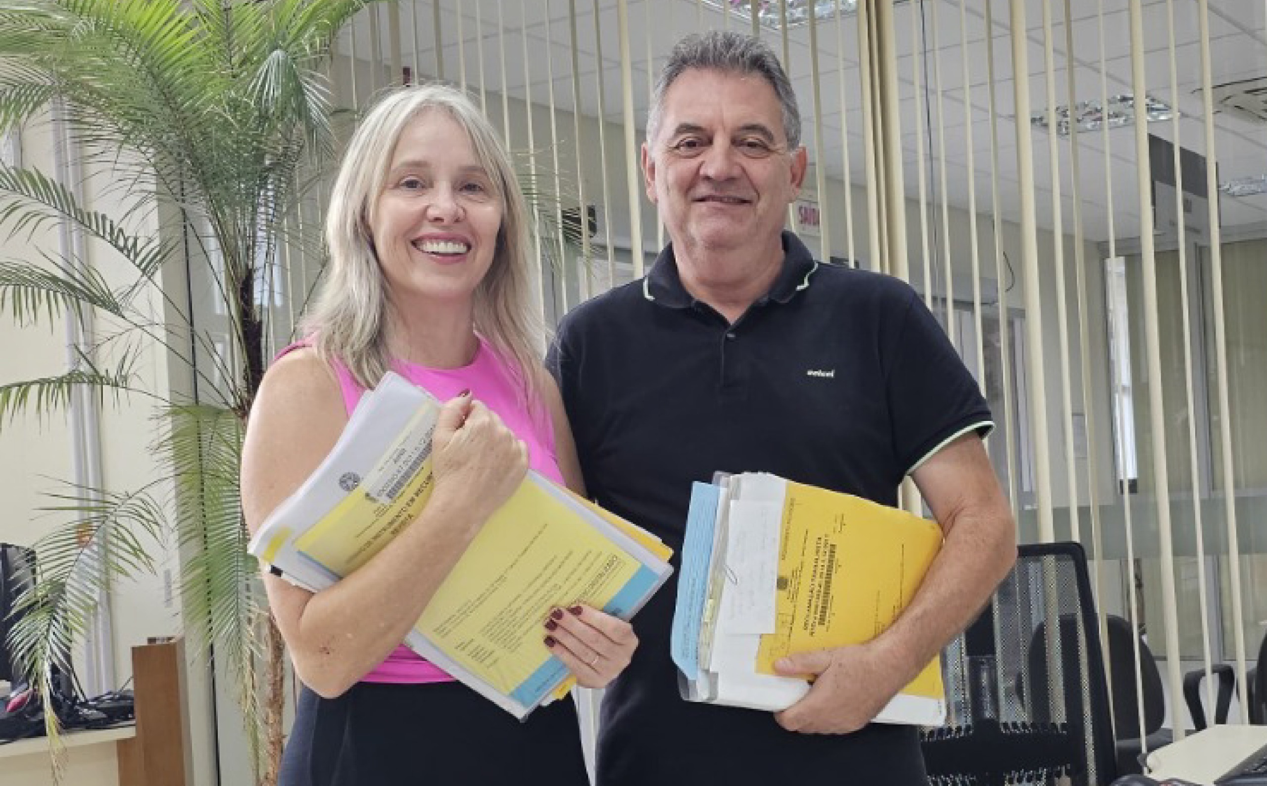 Um homem e uma mulher posam para foto sorridentes, segurando processos embaixo dos braços