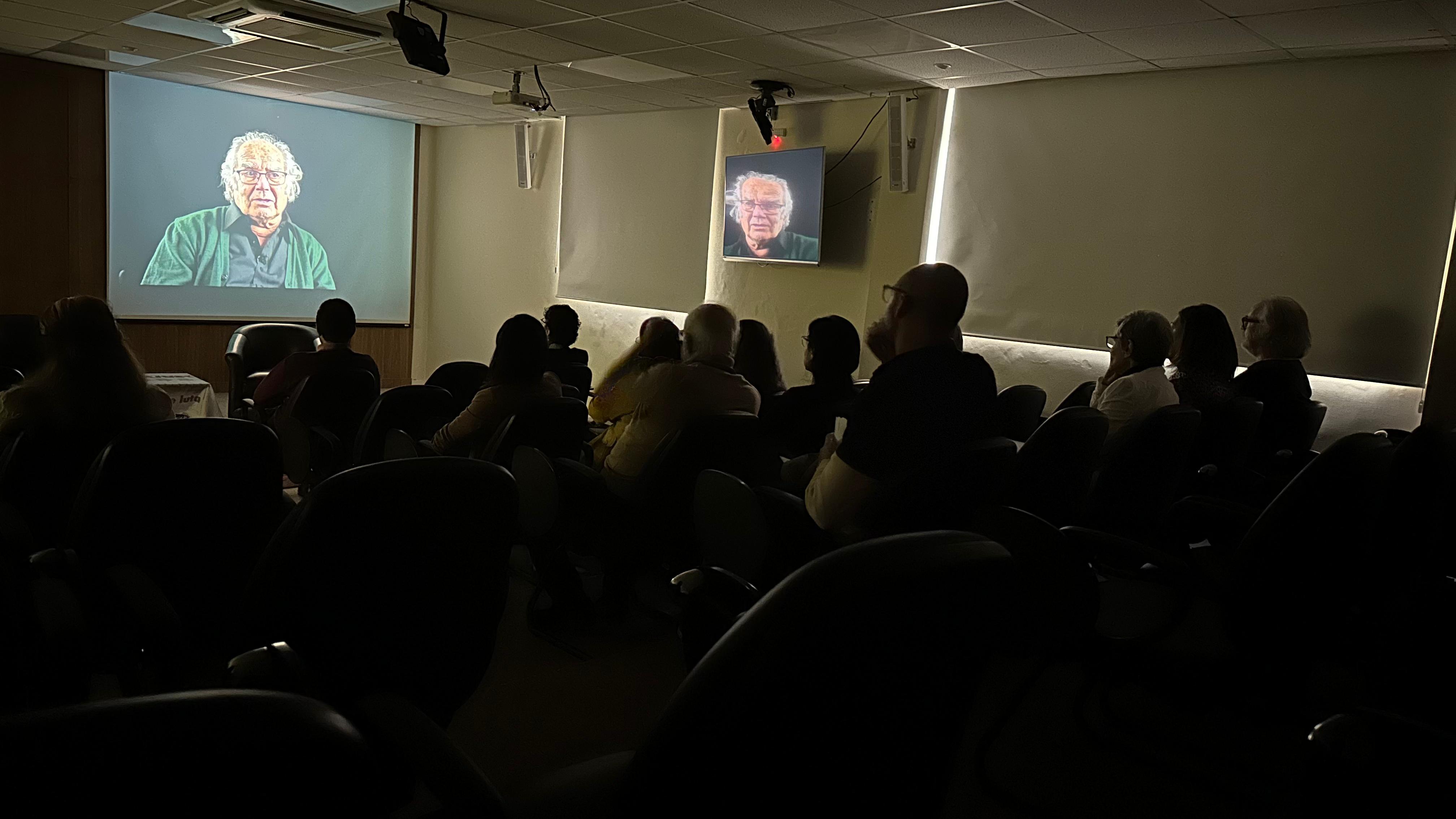 Fotografia de um auditório com as luzes apagadas, no qual o público, sentado, assiste a um documentário projetado sobre uma tela branca localizada na frente do palco.