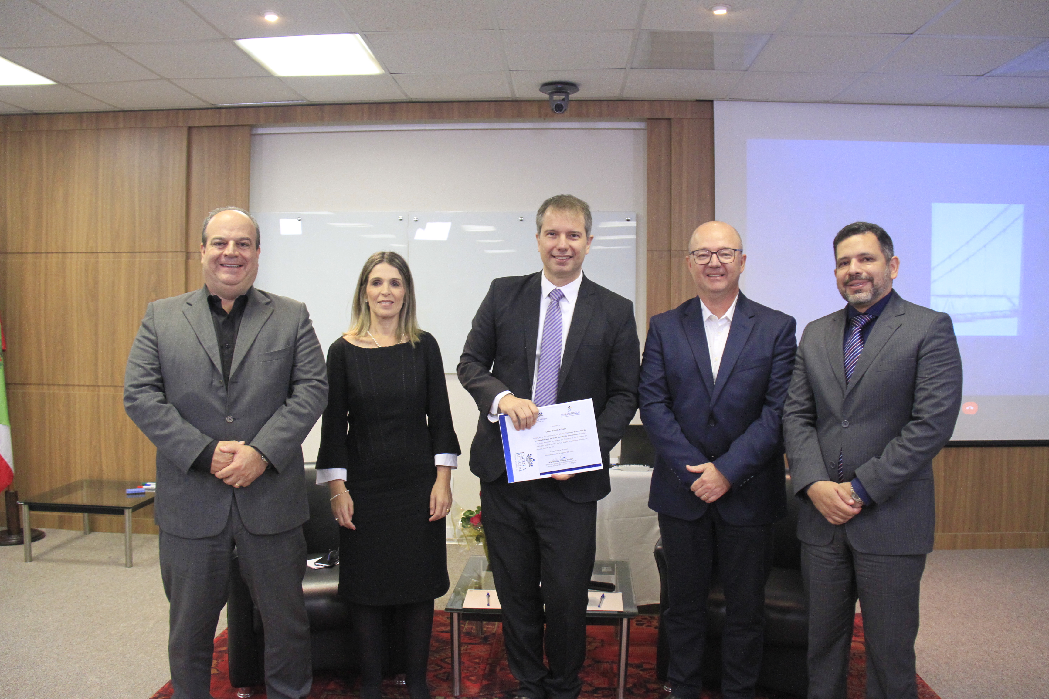 Quatro homens de terno e uma mulher de vestido preto posam para a foto em um auditório. O homem do meio segura um certificado.