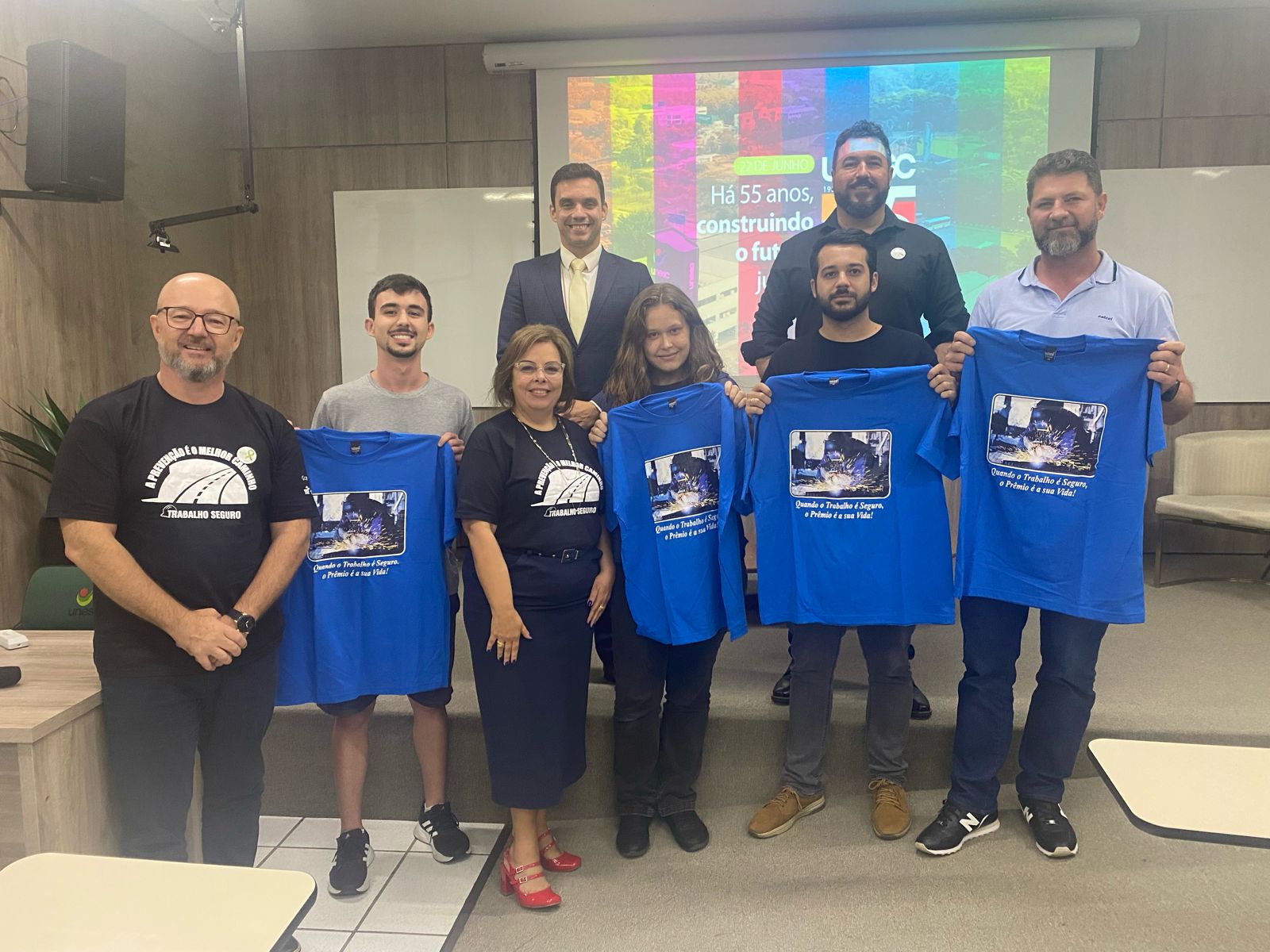 Grupo de pessoas diversas sobre um palco, com alguns segurando camisetas azuis