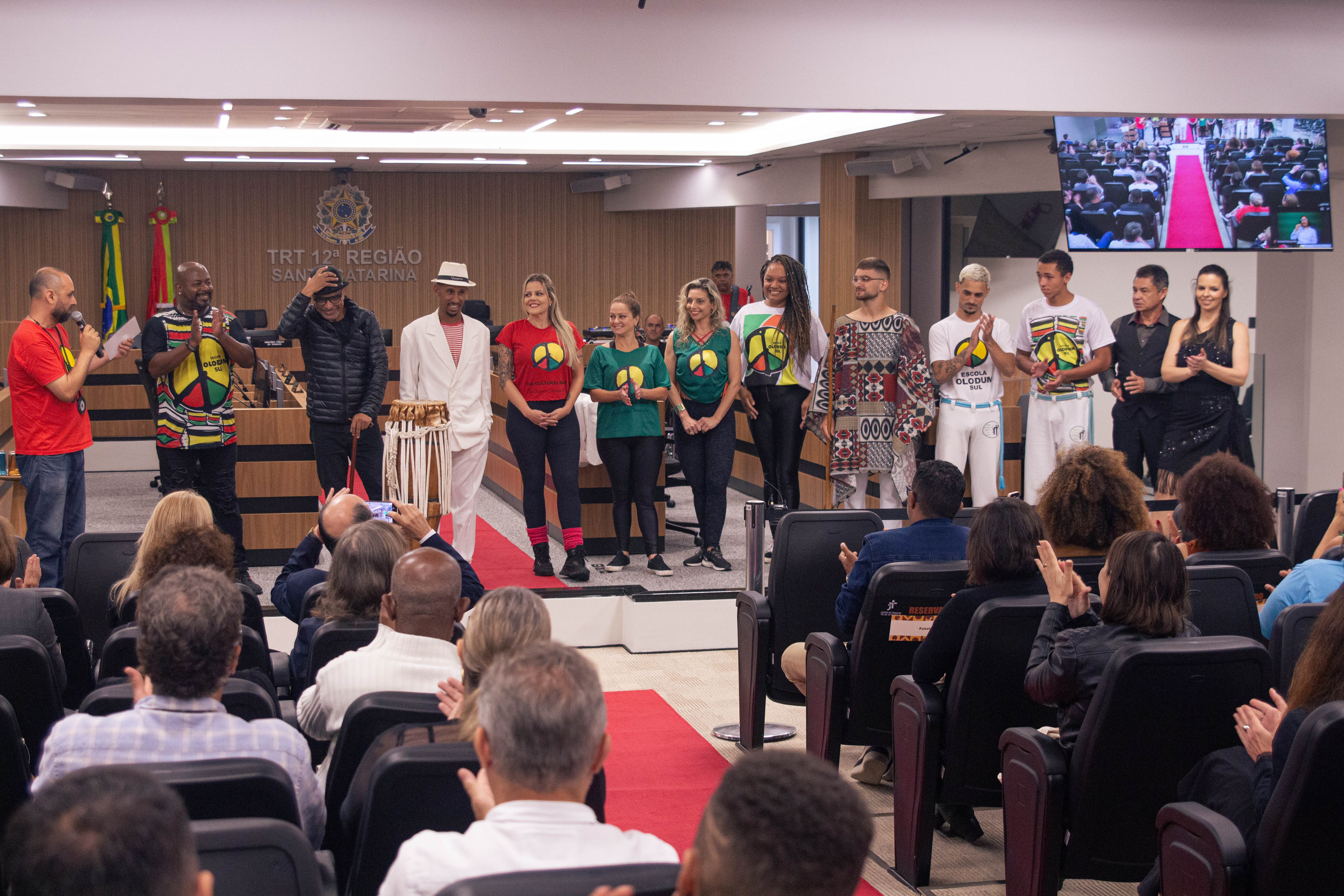 A imagem mostra um grupo de pessoas em pé em frente a uma audiência em um evento formal dentro de uma sala. Algumas pessoas estão vestidas de forma casual, enquanto outras usam trajes tradicionais ou culturais. 