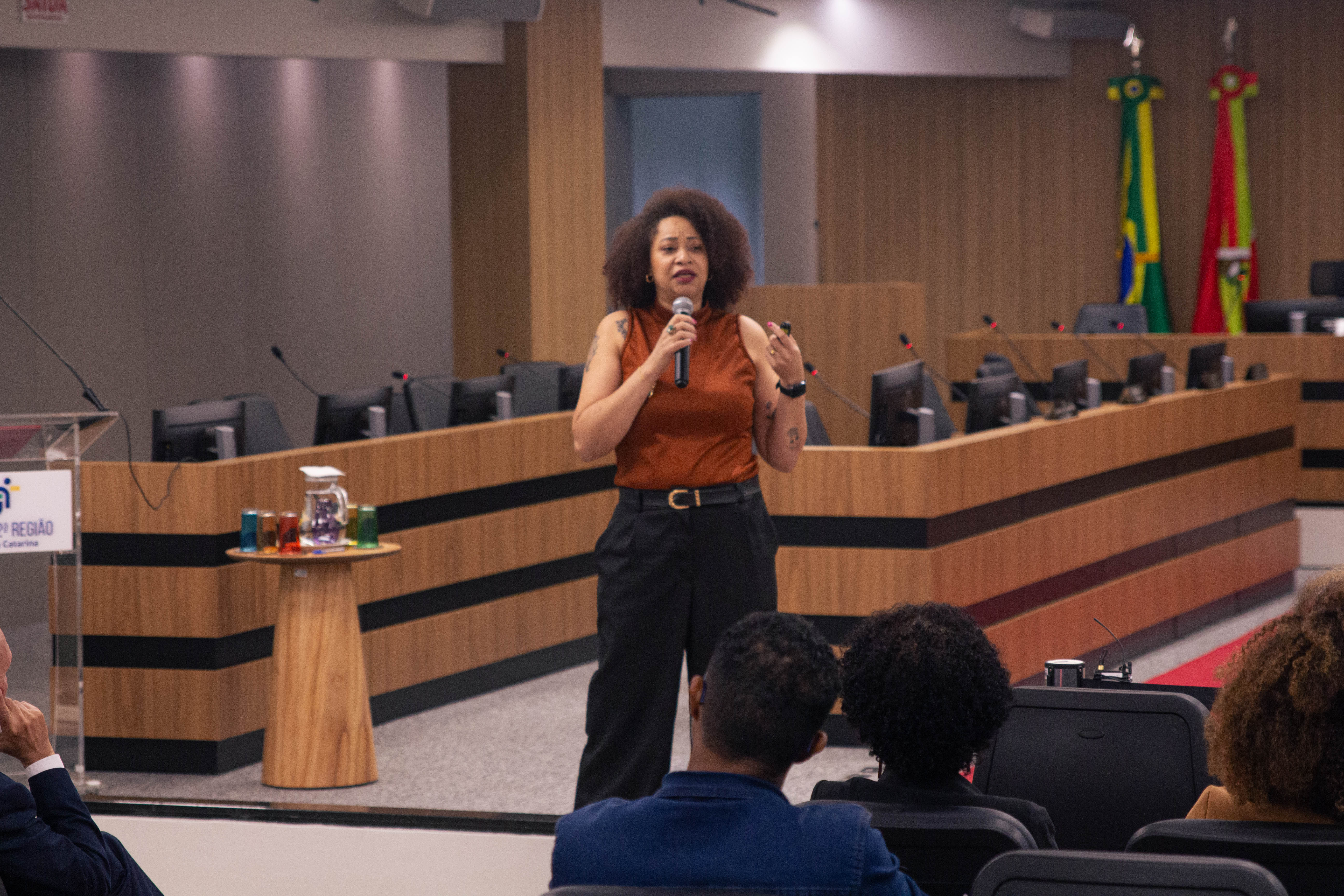 A imagem mostra uma mulher negra de pé, de costas para a câmera, falando para um grupo de pessoas sentadas.