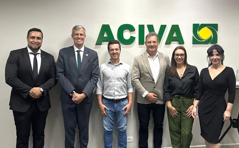 Quatro homens e duas mulheres estão em pé, posando para a foto. Atrás na parede tem a logomarca da empresa Aciva