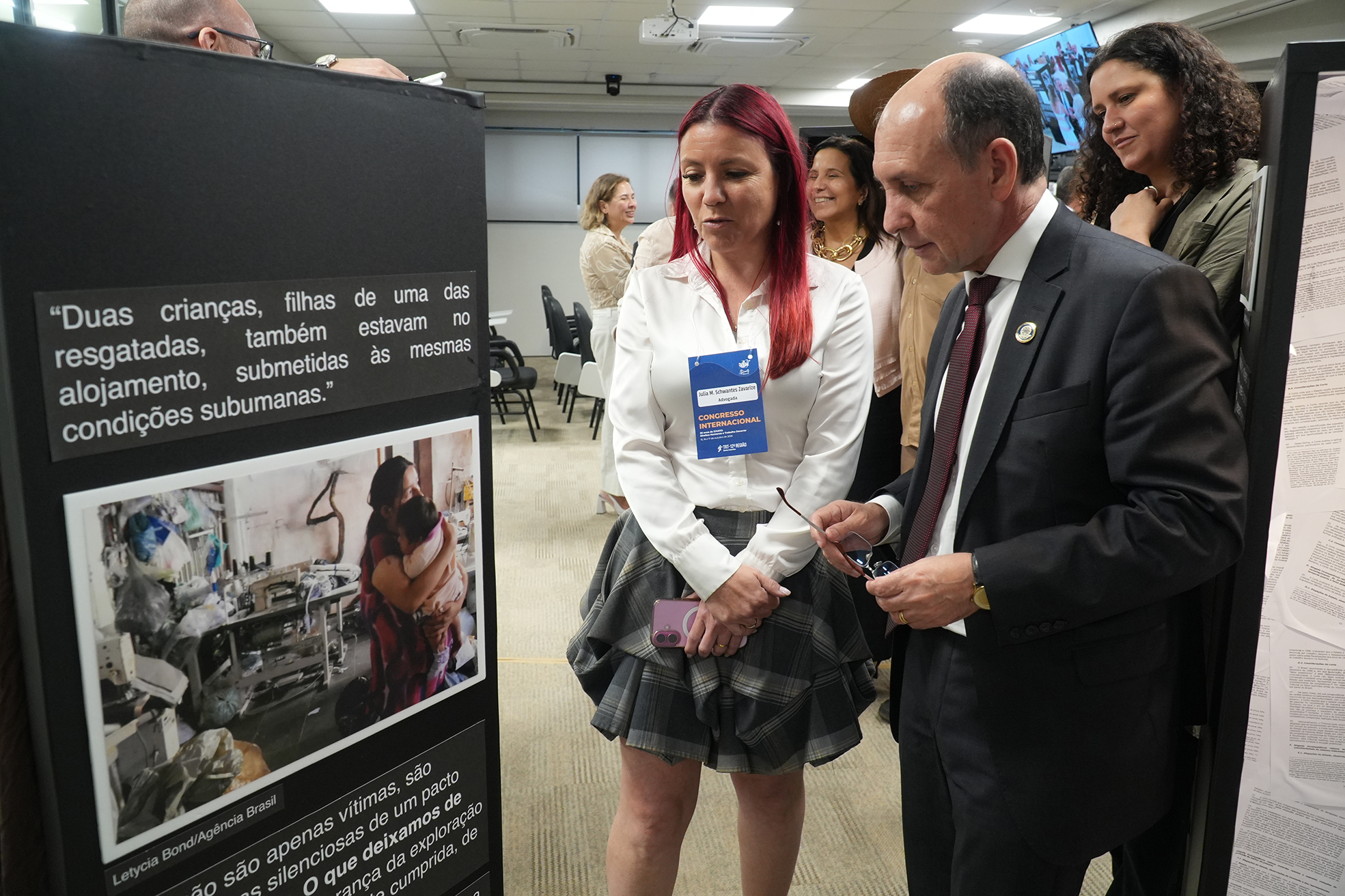 Homem de terno e mulher com roupa social olhando para foto exposta.