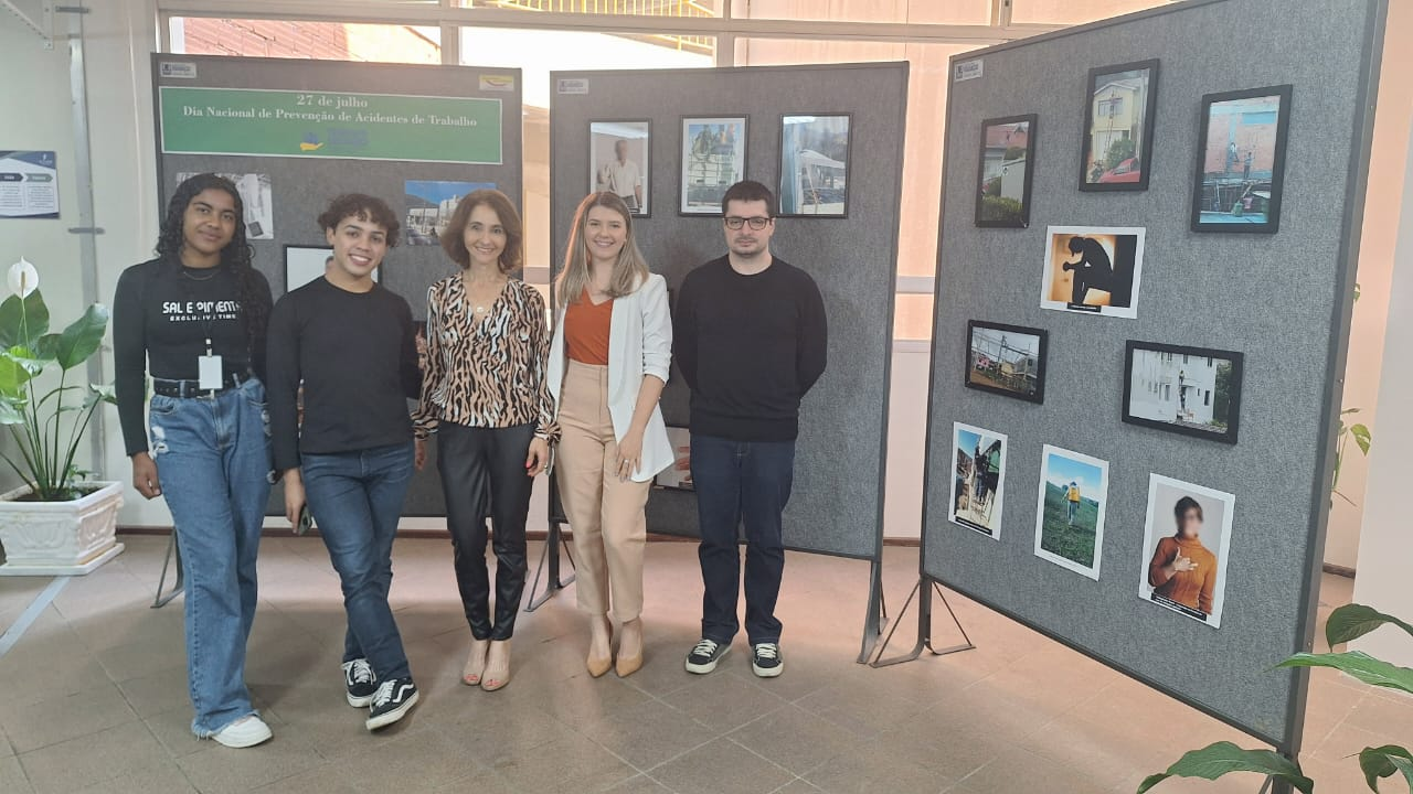 Dois homens e três mulheres estão em pé posando para a foto. Atrás está uma exposição fotográfica