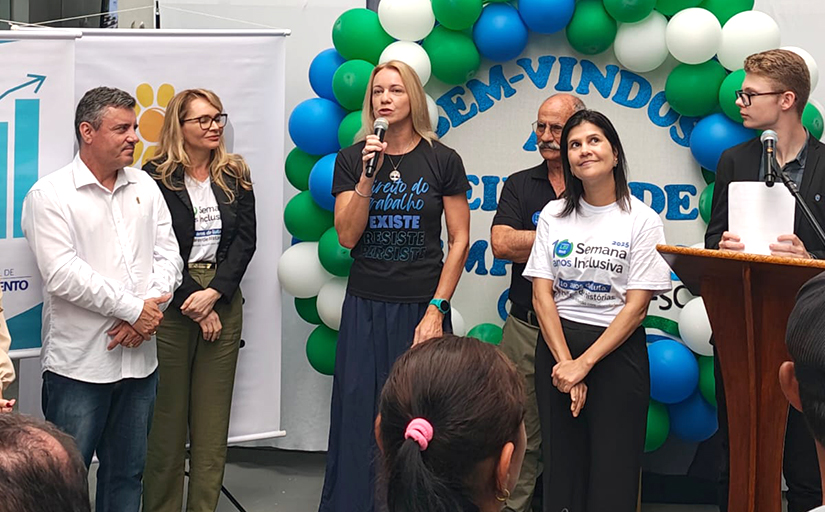 Pessoas em evento com microfone, balões ao fundo e faixas de boas-vindas relacionadas à inclusão e emprego.