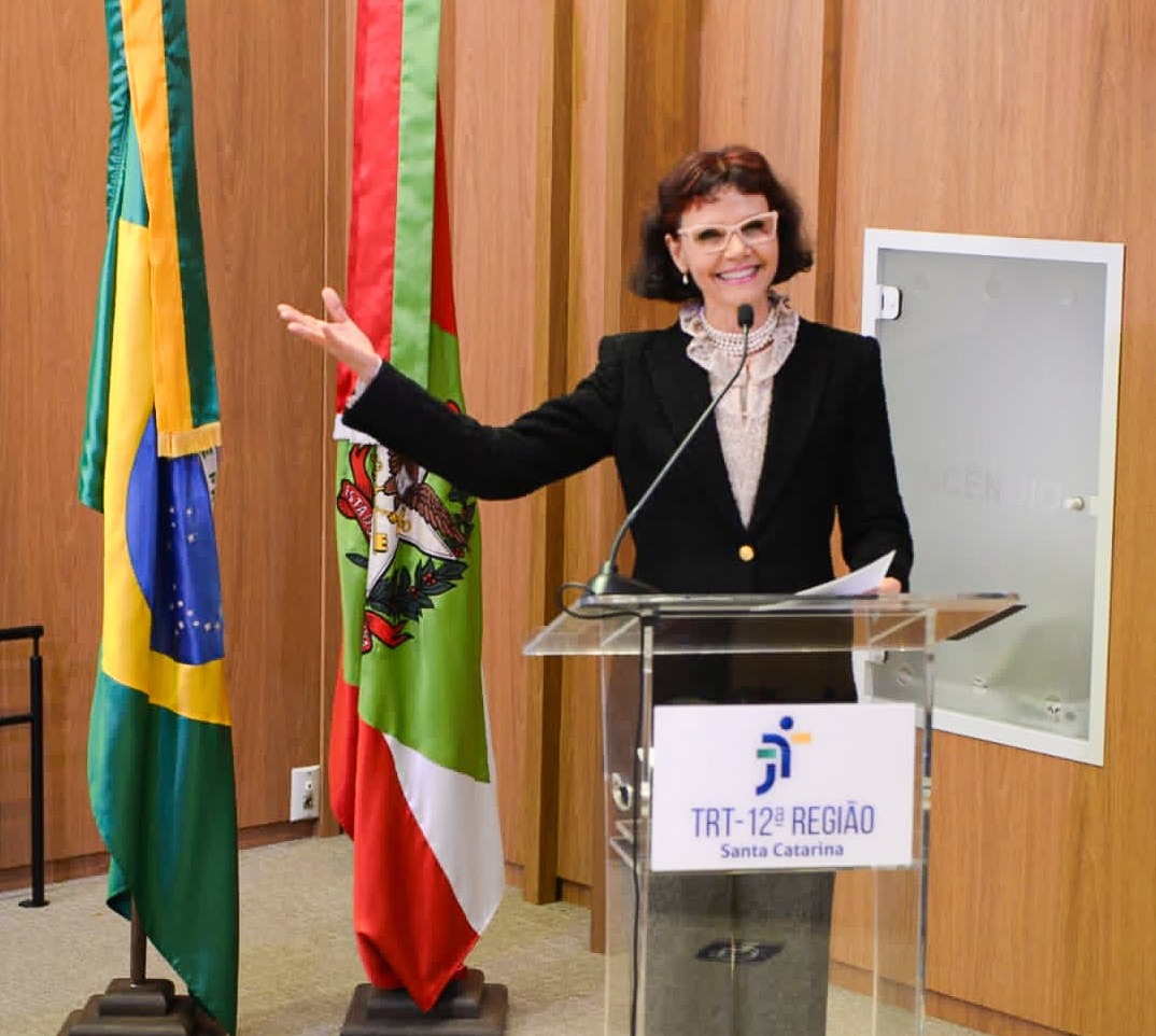 Mulher sorridente discursa atrás de um púlpito. Ela está com um dos braços esticados, tendo bandeiras do Brasil e de SC ao fundo