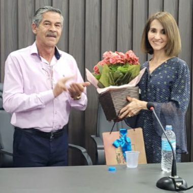 Fotografia de uma mulher sorridente com um arranjo de flores nas mãos. Do lado dela, na esquerda, um homem idoso bate palmas