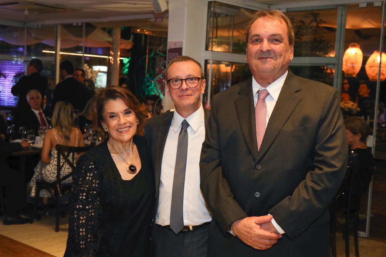 Uma mulher vestida com traje de gala e dois homens de terno posam para a foto, todos sorrindo