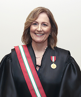 Uma mulher de cabelos castanhos claros, vestindo uma toga e uma faixa está sorrindo para a foto