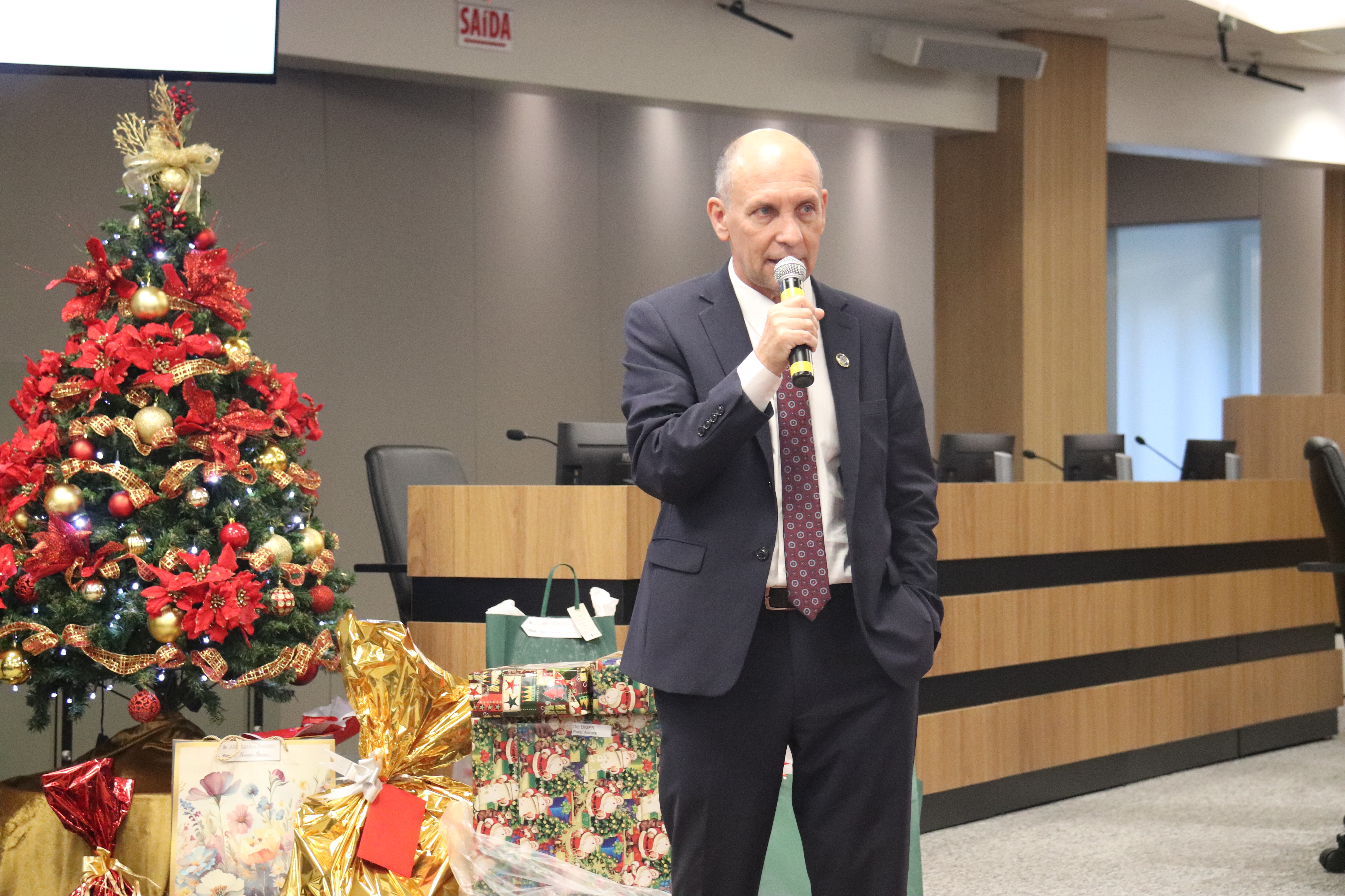 Fotografia de um homem branco de olhos claros, em pé, trajando um terno escuro, camisa branca e gravada bordô. Ele fala ao microfone. À esquerda, há uma árvore de Natal decorada com presentes embaixo.