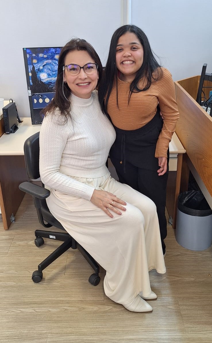 Foto posada de duas mulheres sorrindo abraçadas em um ambiente de escritório. À esquerda está uma mulher branca de cabelo escuro liso, que veste blusa e saia branca, ela usa óculos e está sentada em uma cadeira de escritório, ao lado dela está uma mulher mais nova de pele clara, de cabelo preto liso, que veste uma blusa laranja quadriculada e está de pé.