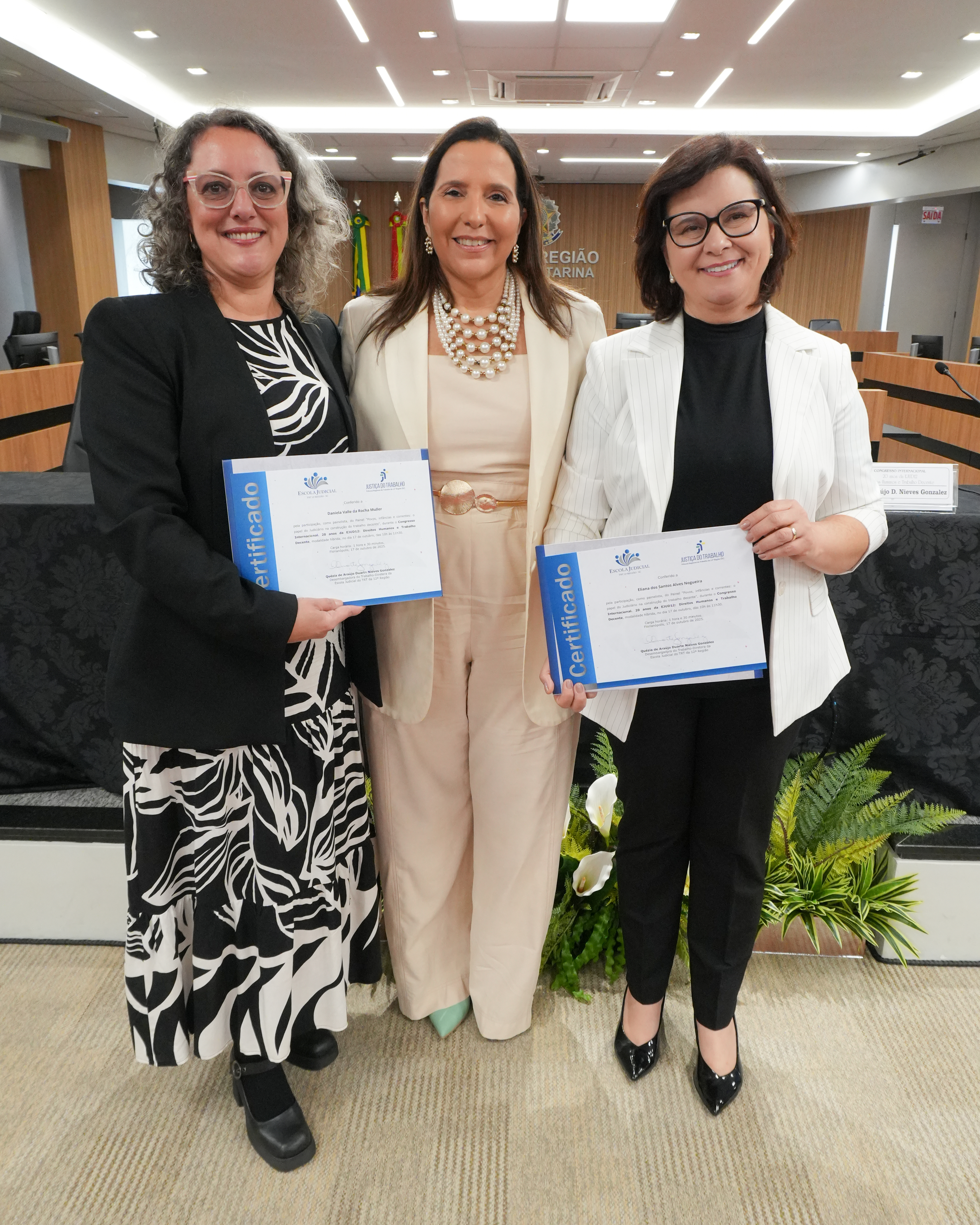 Três mulheres em pé e sorrindo para a foto, lado a lado. Duas delas estão segurando um certificado
